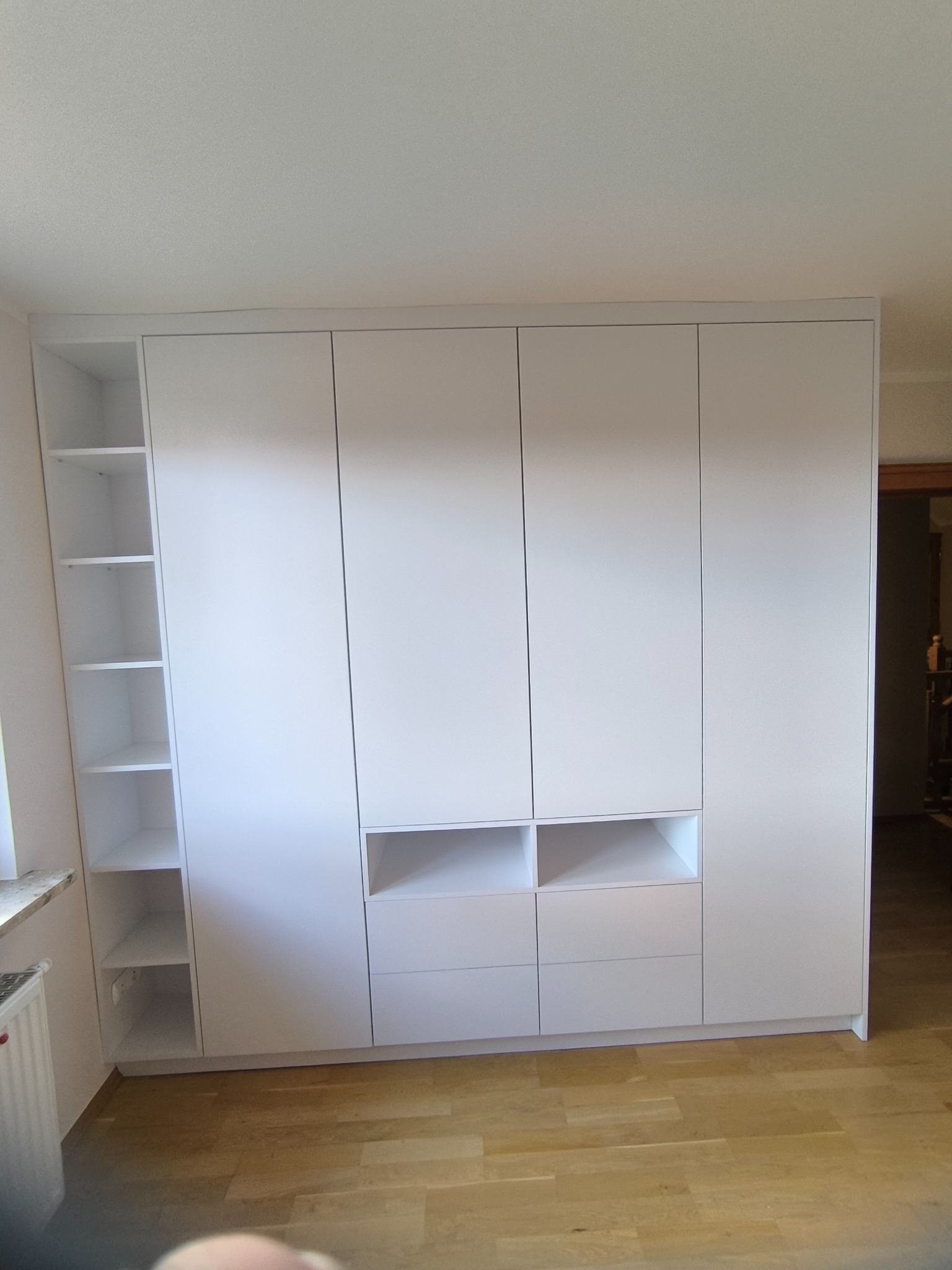 Biała szafa wnękowa z półkami i szufladami, idealnie dopasowana do wnętrza. Minimalistyczny design, drewniana podłoga, jasne ściany.