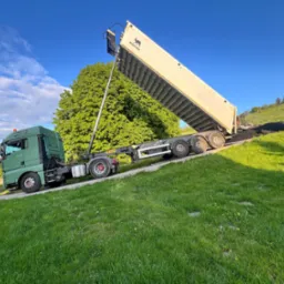 Zielony samochód ciężarowy z otwartą naczepą wysypuje asfalt na zielonym terenie. W tle drzewa i pagórki pod błękitnym niebem.
