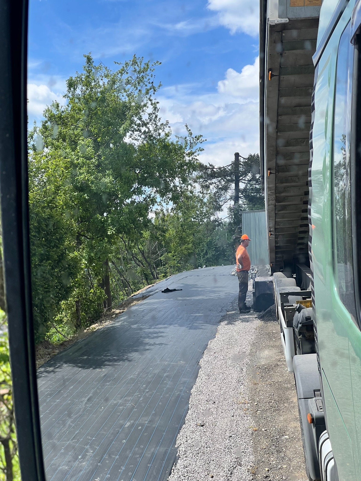 Układanie geowłókniny na drodze gruntowej, widok z kabiny ciężarówki. Pracownik w pomarańczowej czapce nadzoruje proces utwardzania podłoża.