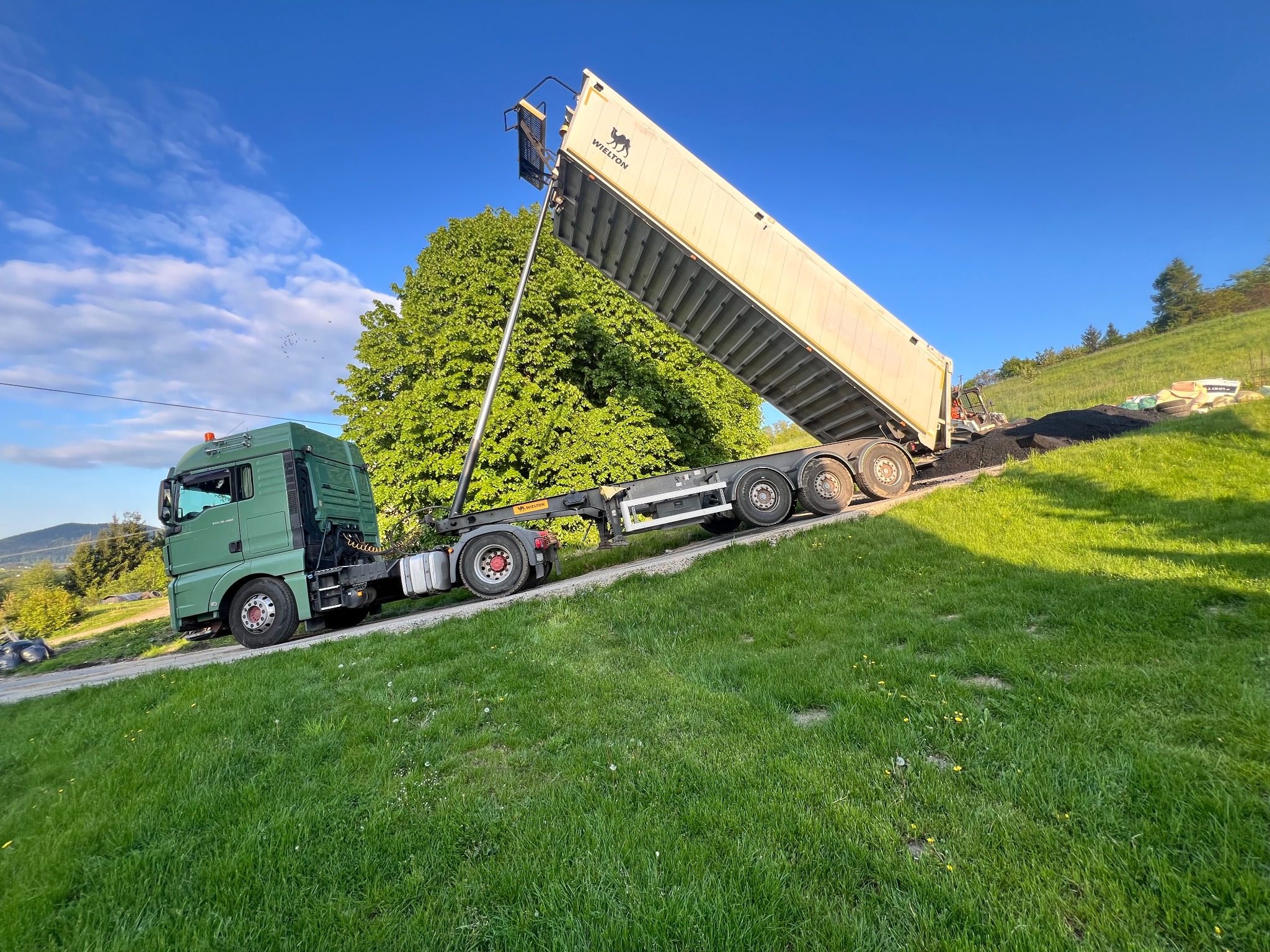 Zielony samochód ciężarowy z otwartą naczepą wysypuje asfalt na zielonym terenie. W tle drzewa i pagórki pod błękitnym niebem.