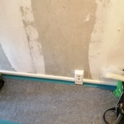 EL Mar MARCIN SOBCZAK - Białe gniazdko elektryczne zamontowane przy podłodze w pomieszczeniu w trakcie remontu. Widoczne ślady po szpachlowaniu na ścianie.