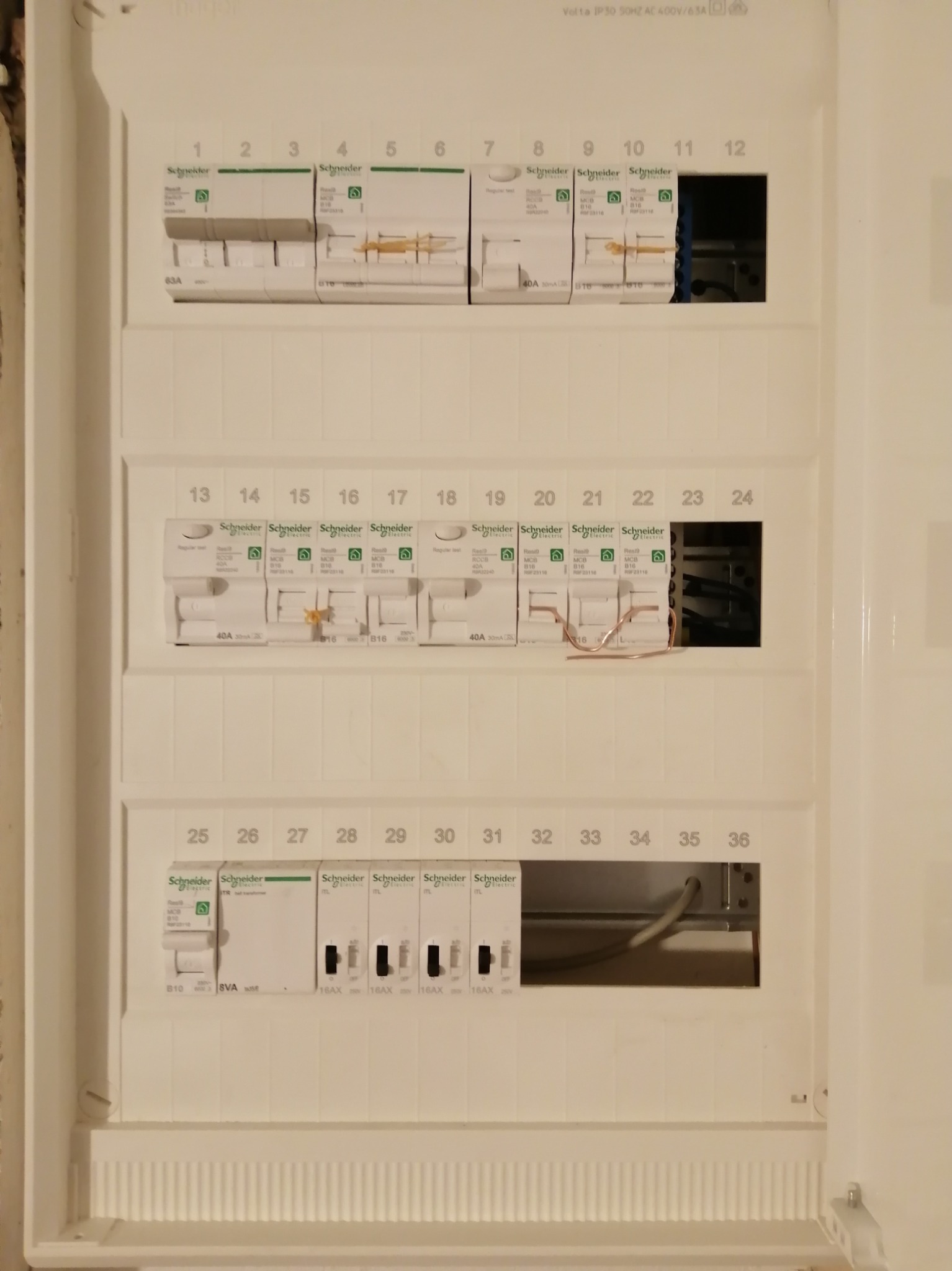 Rozdzielnia natynkowa Schneider z bezpiecznikami, niektóre obwody zabezpieczone prowizorycznie przed włączeniem napięcia. Widoczne kable i numery obwodów.