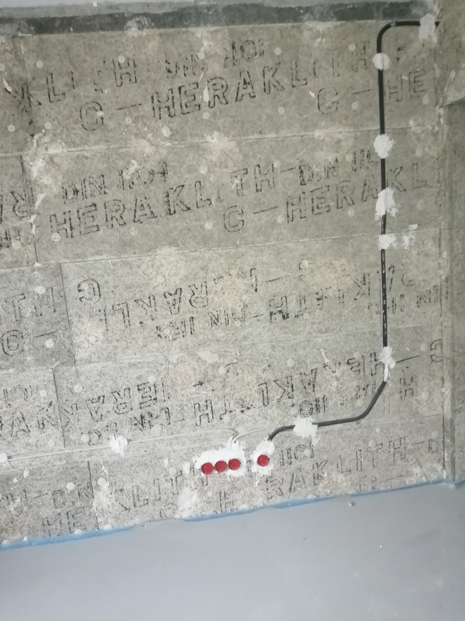 Czarny przewód elektryczny przymocowany do płyty Heraklith, z widocznymi czerwonymi końcówkami kabli. Instalacja w trakcie montażu.