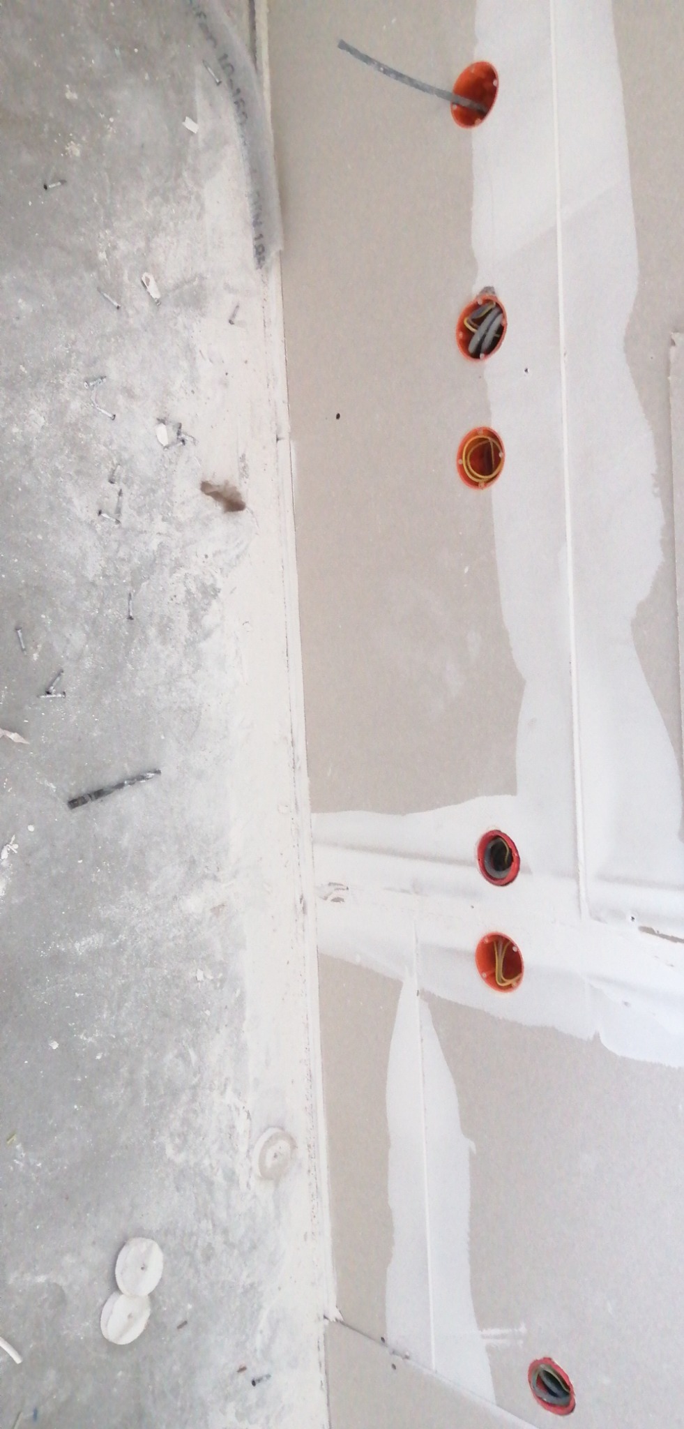Montaż instalacji elektrycznej w ścianie z płyt gipsowo-kartonowych. Widoczne pomarańczowe puszki instalacyjne z przewodami i fragment nieuprzątniętej podłogi.