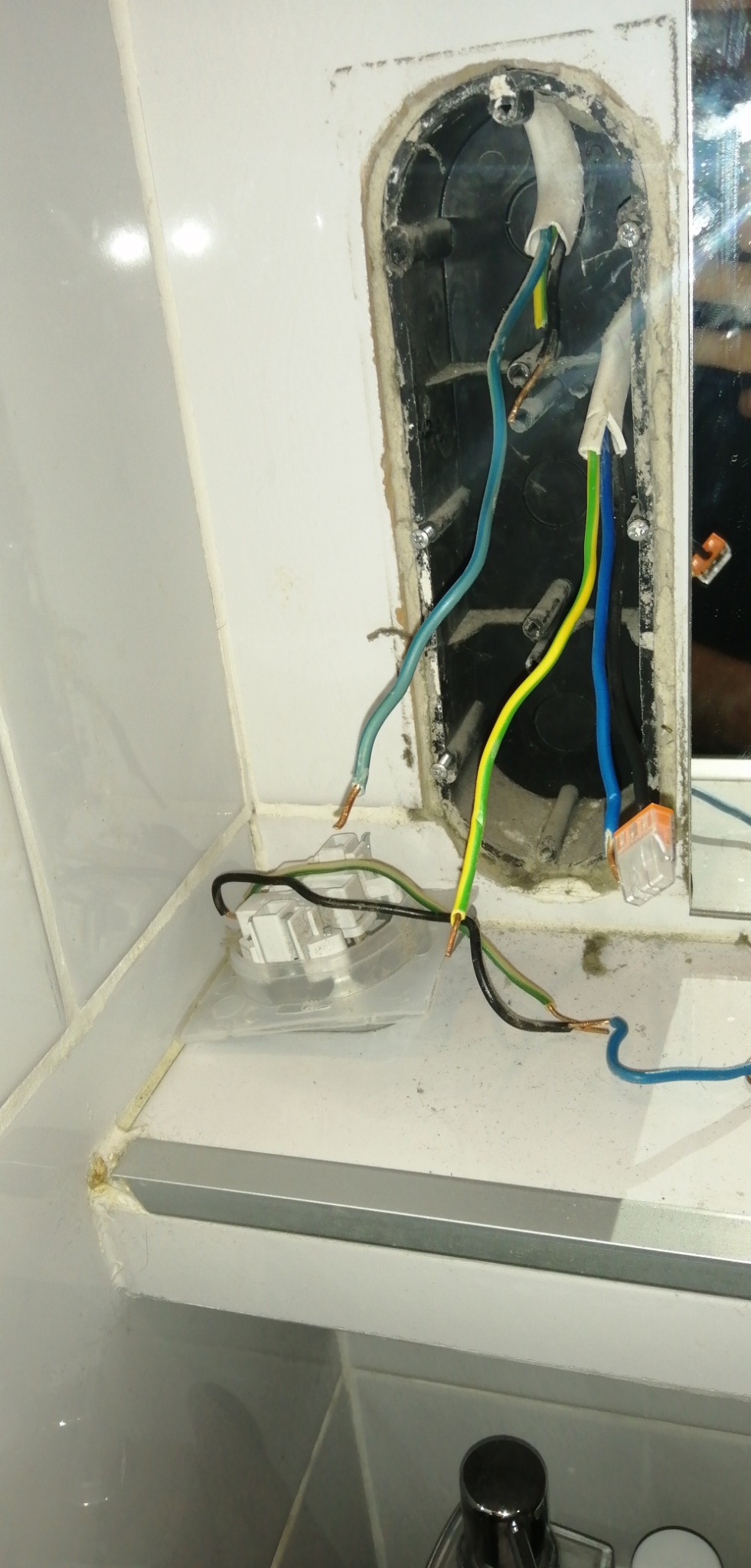 Odsłonięta puszka elektryczna w łazience z wystającymi kablami, widoczne złącza WAGO. Brak oprawy, płytki ceramiczne w tle.