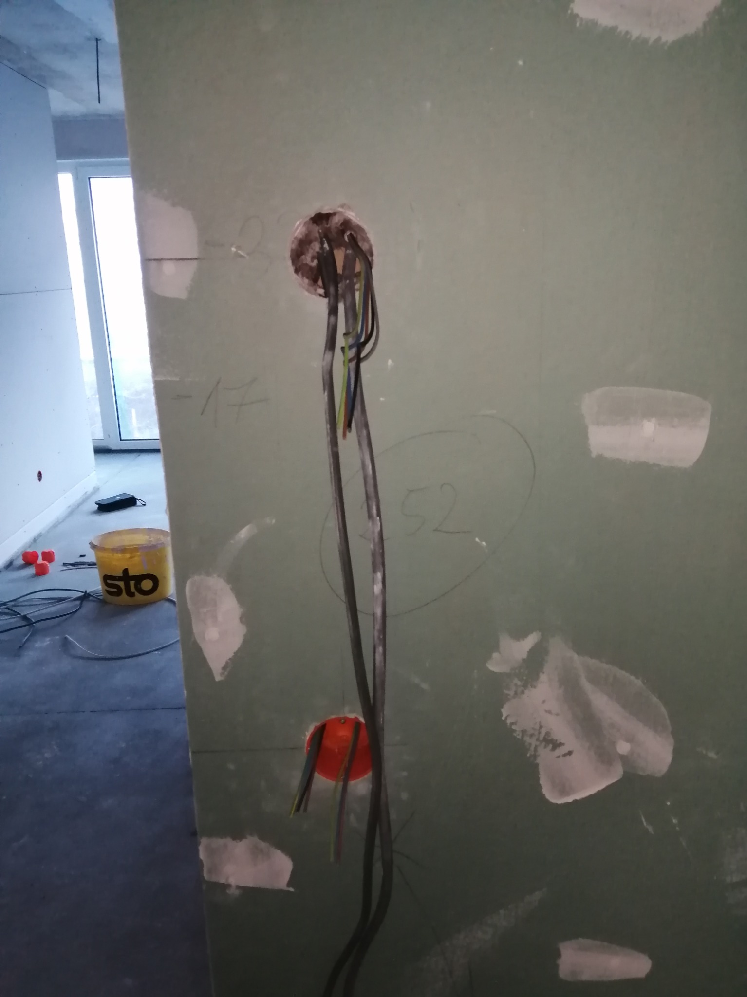 Ściana z płyty gipsowej z dwoma otworami pod instalację elektryczną, widoczne kable i puszki. Częściowo zaszpachlowana, na podłodze stoi wiadro.