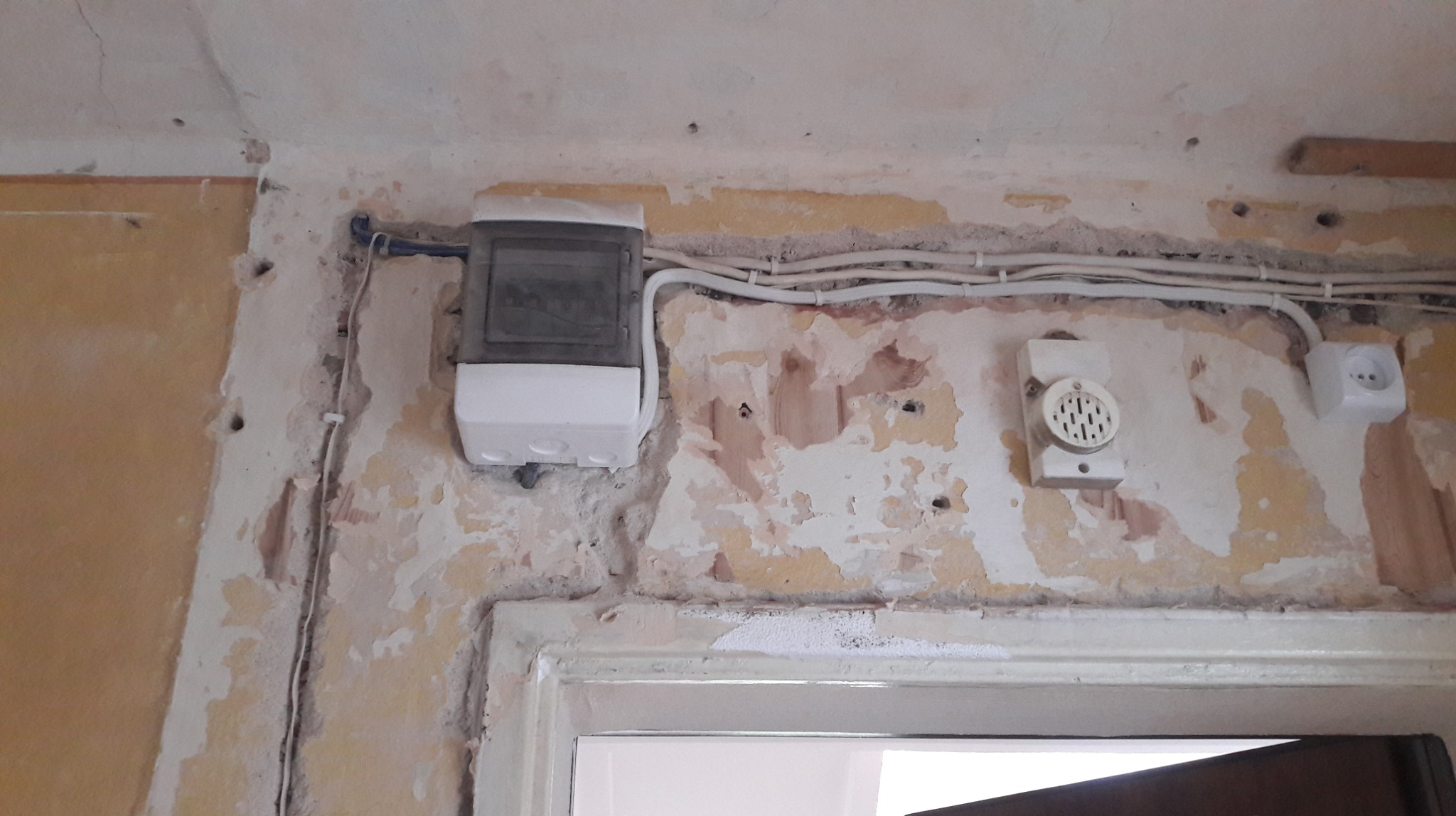 Biała skrzynka elektryczna, przewody, gniazdko i głośnik na starej, częściowo obdrapanej ścianie nad futryną drzwi. Widoczne ślady po instalacji.