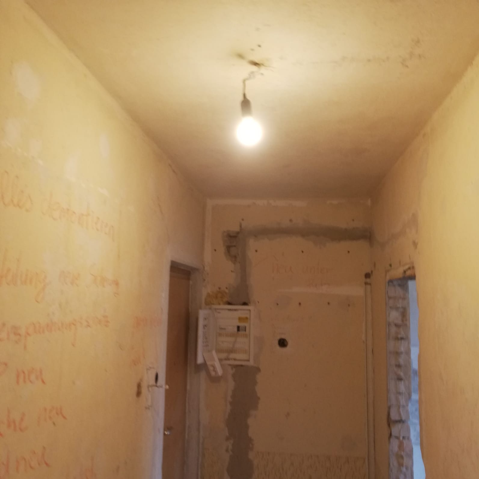 Wnętrze w trakcie remontu: widoczna skrzynka elektryczna na ścianie, surowe ściany z napisami, wisząca żarówka. Prace przygotowawcze do instalacji.