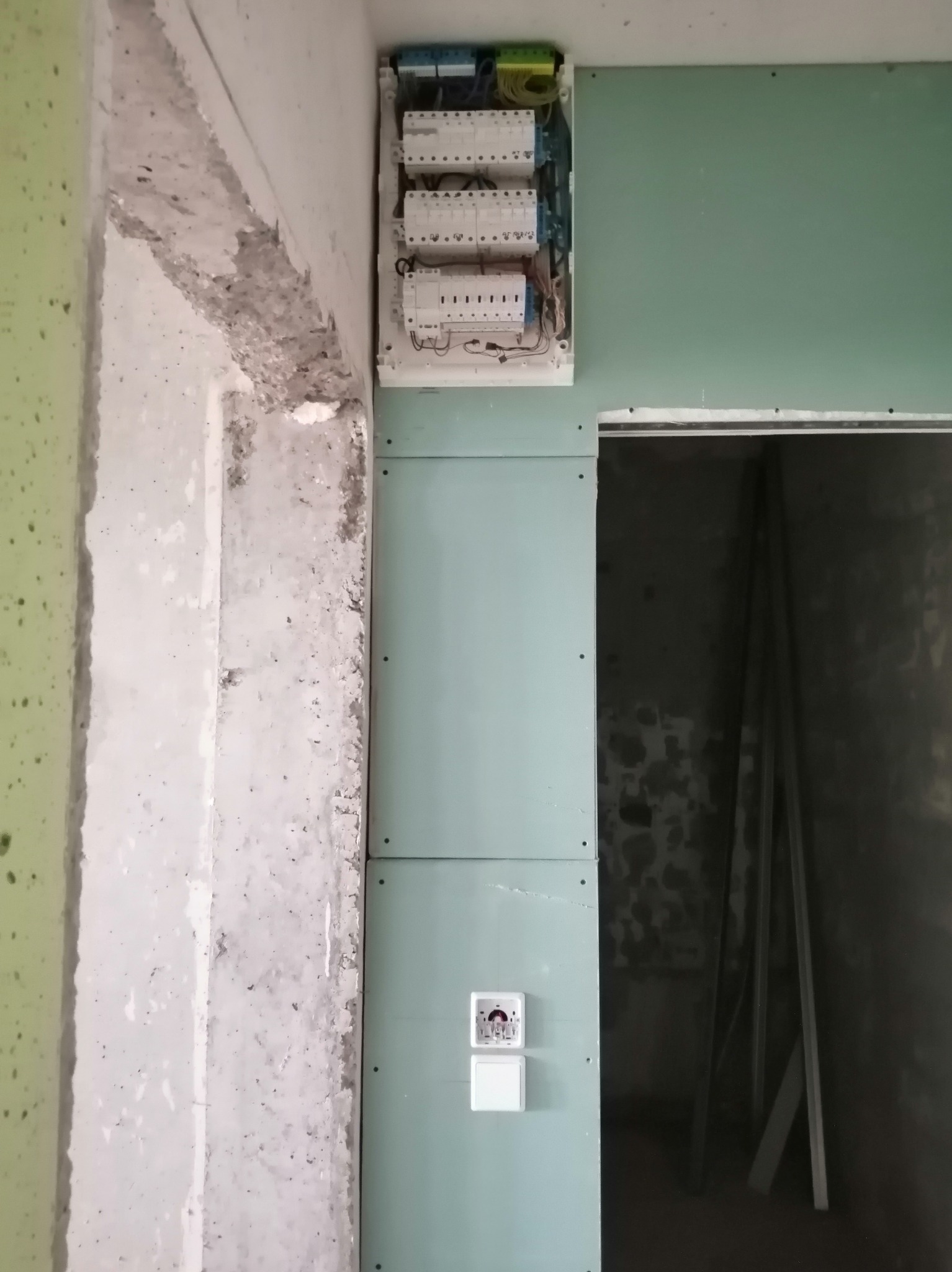 Rozdzielnica elektryczna zamontowana na ścianie z płyt gipsowo-kartonowych w surowym wnętrzu, widoczne kable i zabezpieczenia. Etap prac wykończeniowych.