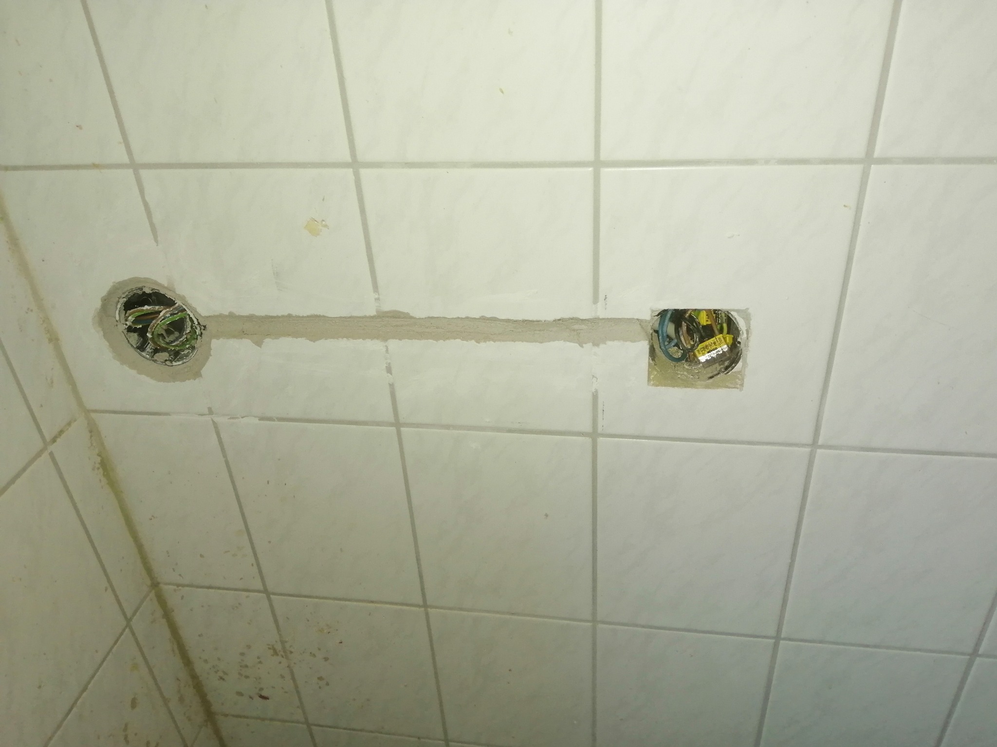 Białe płytki, bruzda z przewodami i dwa otwory pod puszki elektryczne. Widoczne kable w kolorach: zielonym, niebieskim i żółtym.  Przewody w ścianie.