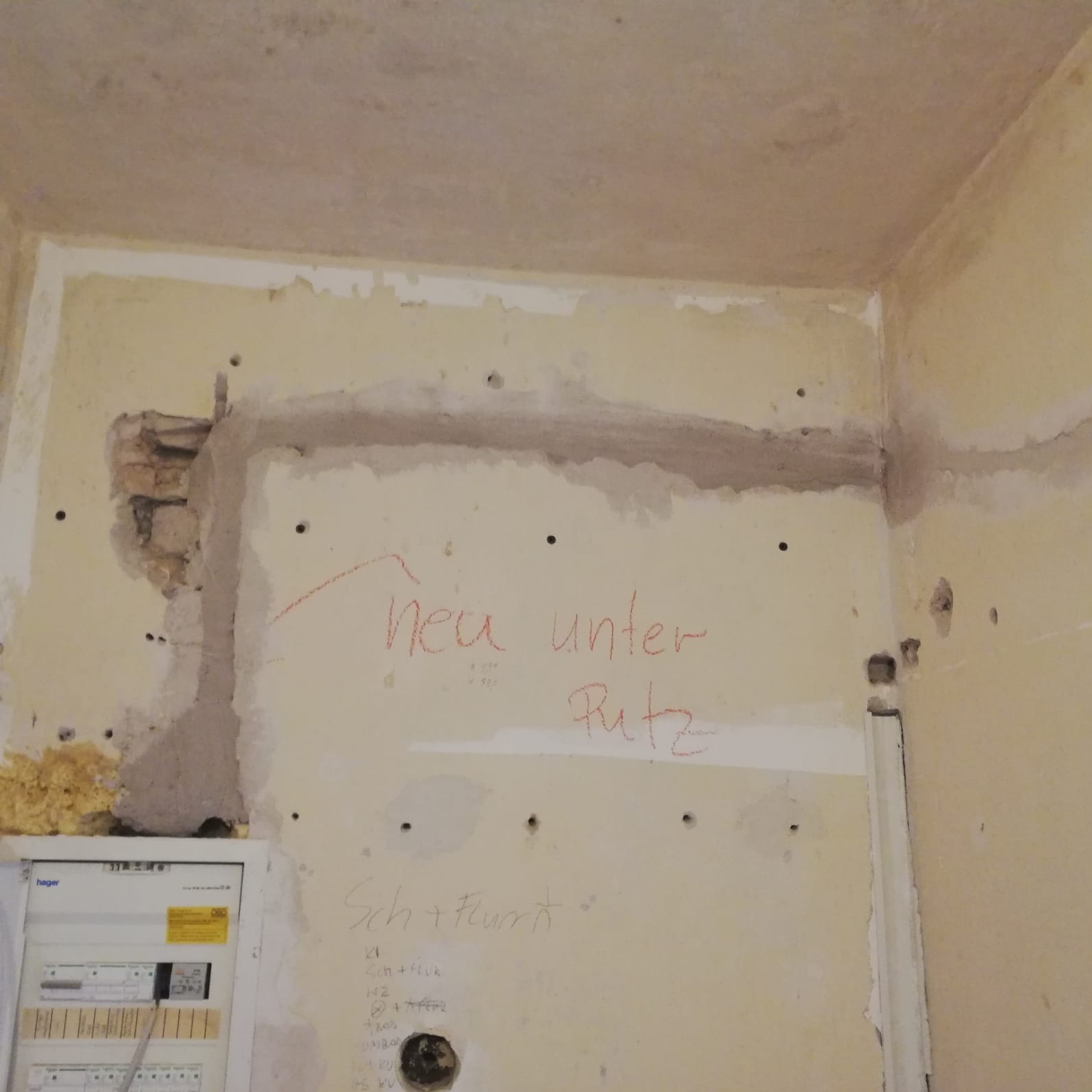 Ściana w trakcie remontu z widocznymi śladami po instalacjach, otwory, napisy 'neu unter Putz' i tablica elektryczna Hager w lewym dolnym rogu.