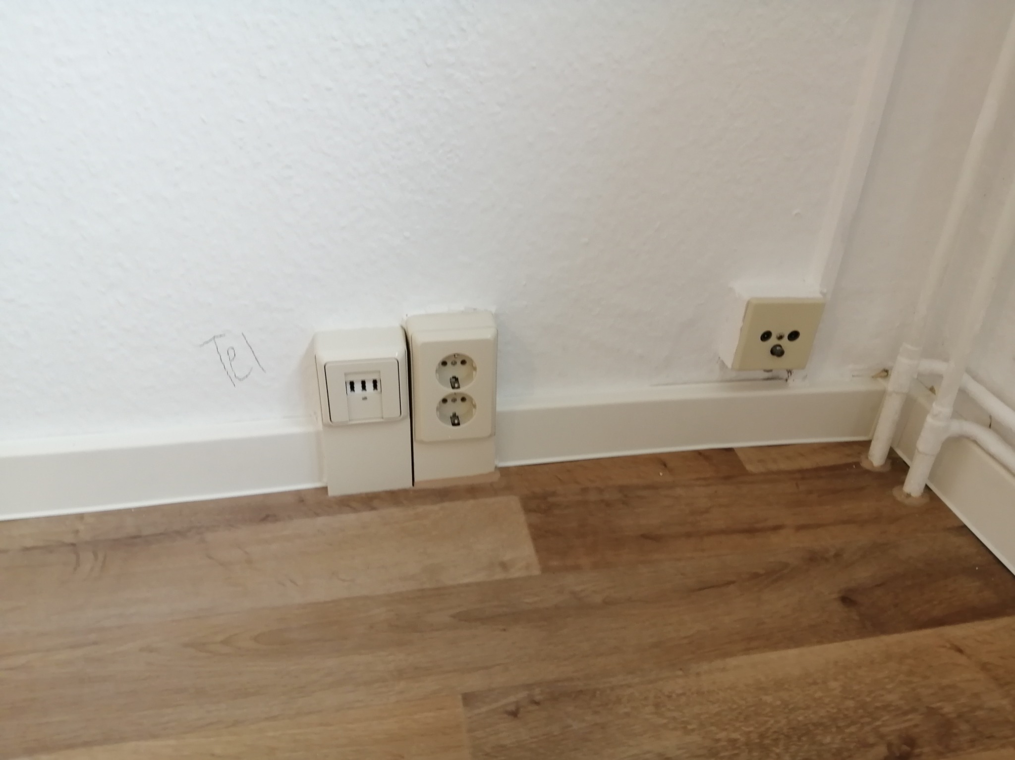 Białe gniazdka elektryczne i telefoniczne zamontowane przy podłodze z paneli. Na ścianie widoczny napis 'Tel'.