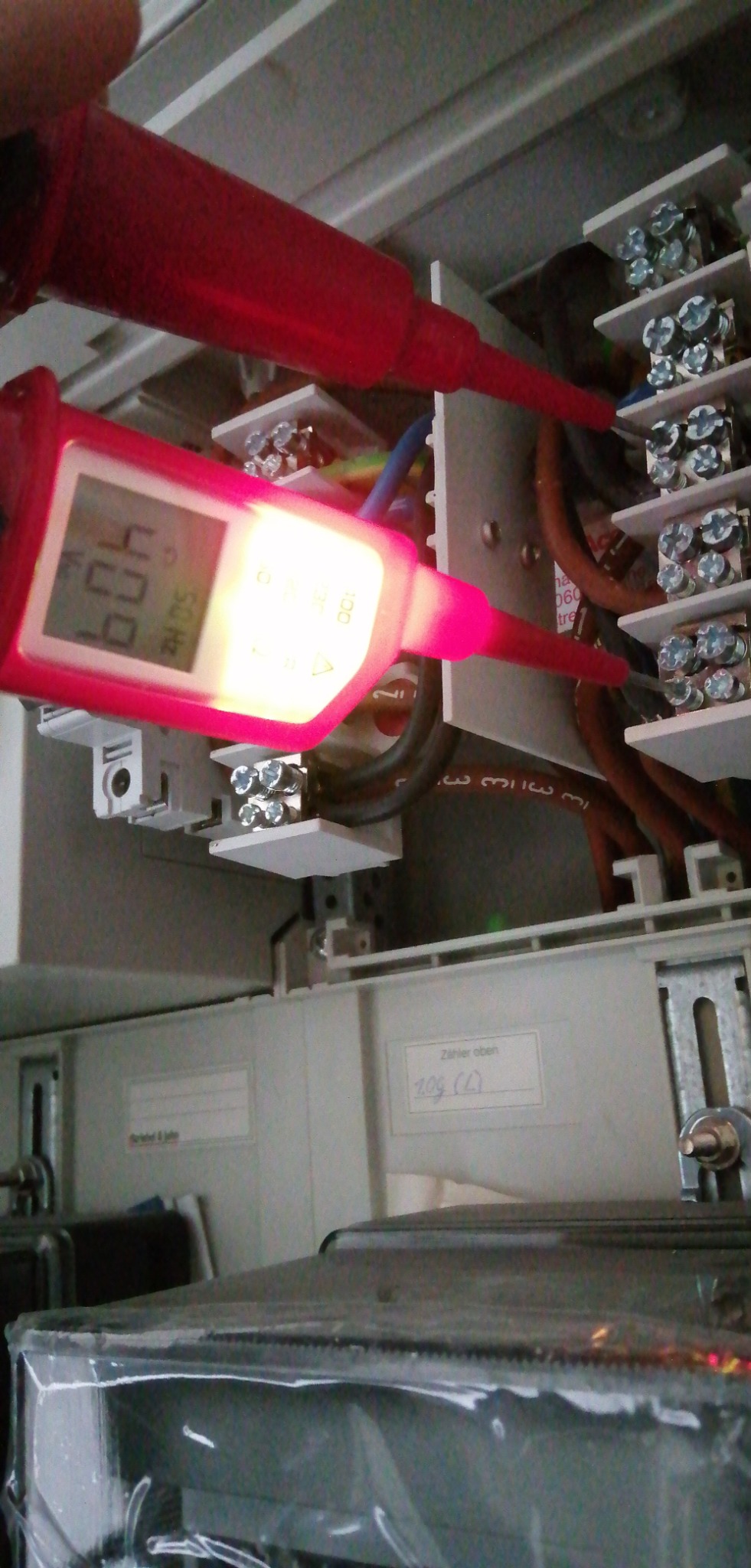 Czerwony miernik napięcia wskazuje 409V na listwie zaciskowej w szarej skrzynce elektrycznej. Widoczne kable i śruby mocujące.