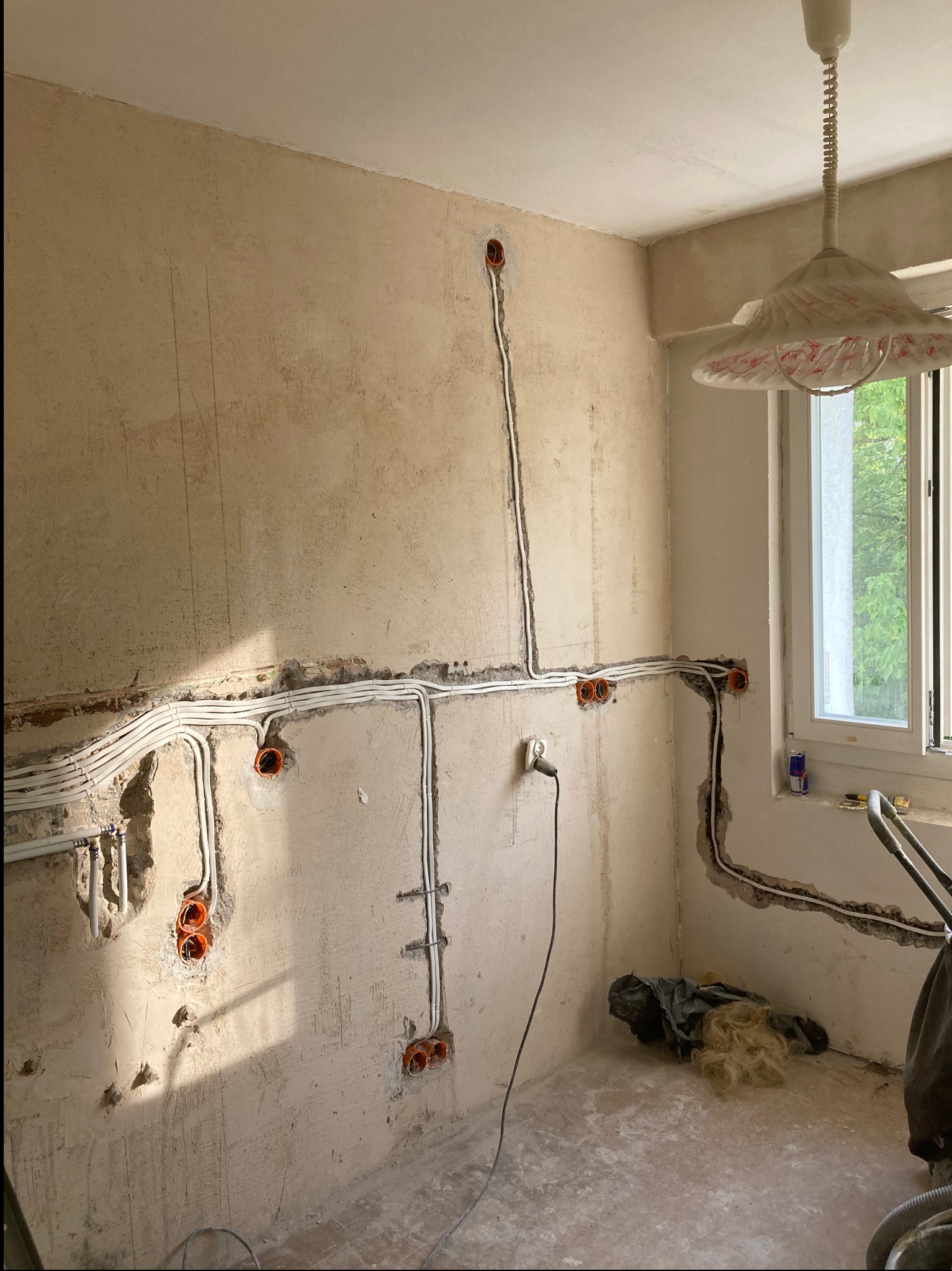 Ściana w trakcie montażu instalacji elektrycznej: kable w bruzdach, pomarańczowe puszki. Widoczne narzędzia i okno z widokiem na zieleń. Wnętrze w trakcie remontu.