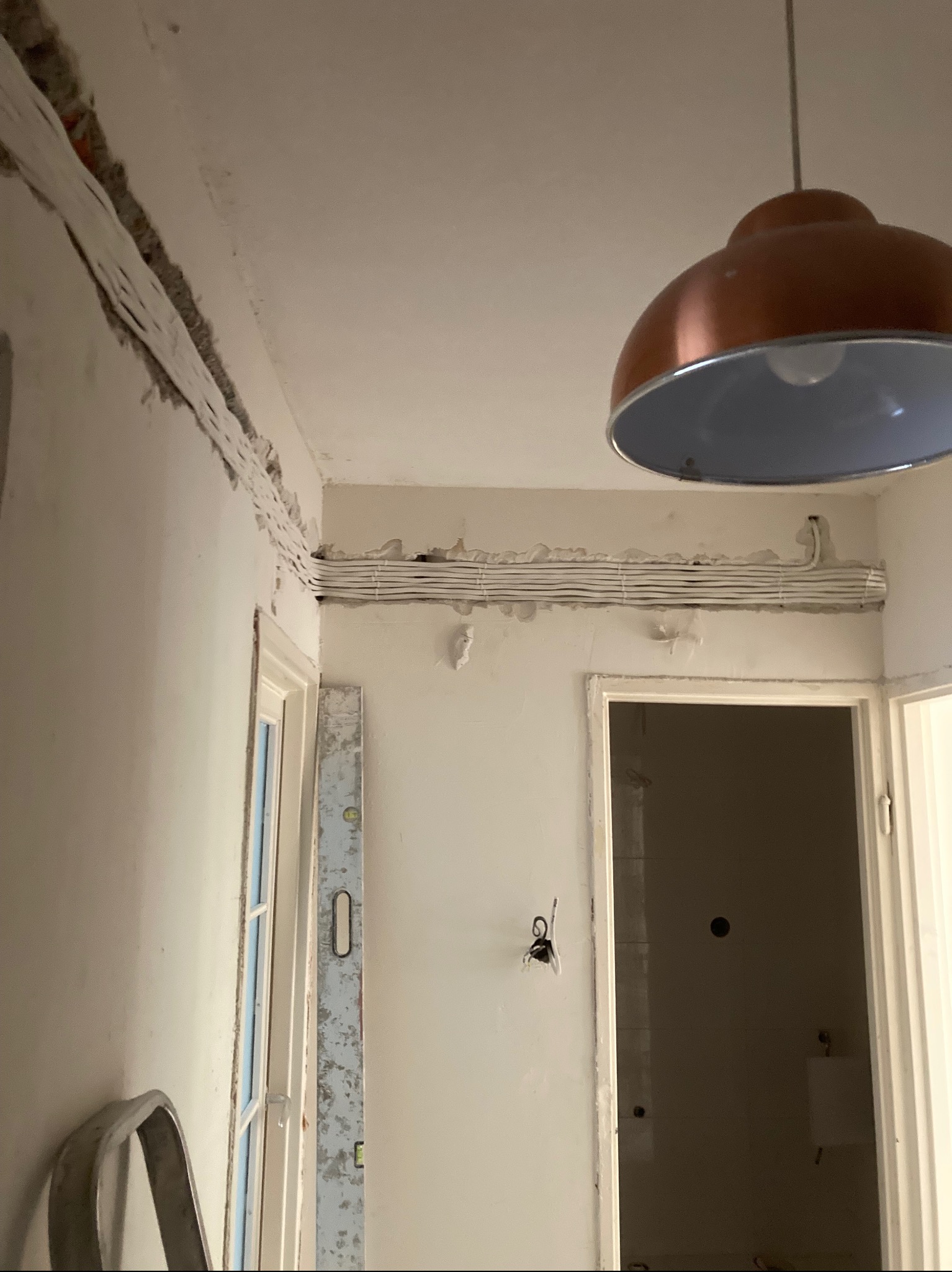 Białe kable ułożone w bruździe na ścianie, widoczna miedziana lampa wisząca i poziomica oparta o futrynę drzwi w trakcie prac remontowych.