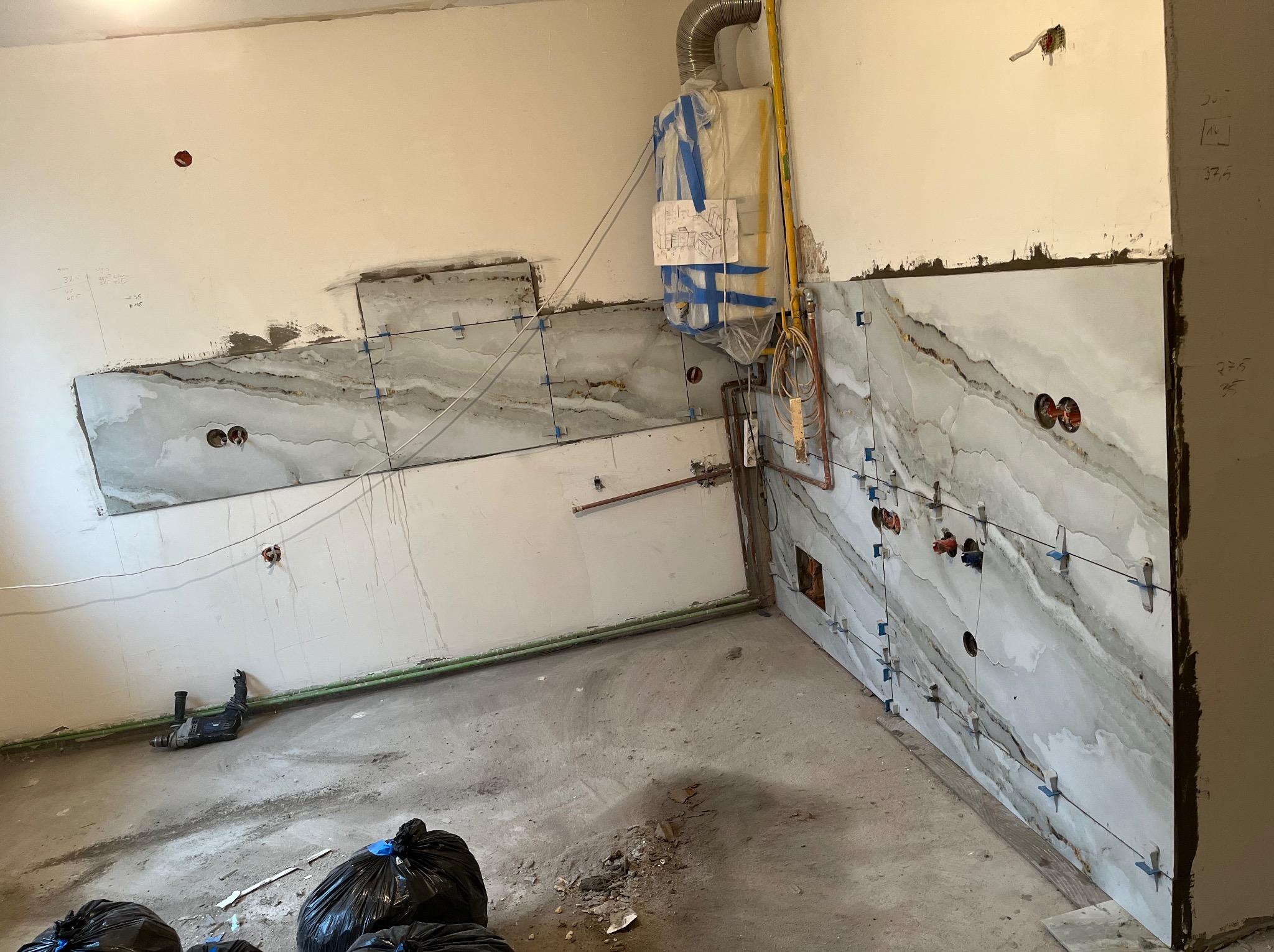 Wnętrze w trakcie układania płytek imitujących marmur. Widoczne krzyżyki glazurnicze, rury i zabezpieczony piec gazowy. Na podłodze gruz i worki na śmieci.