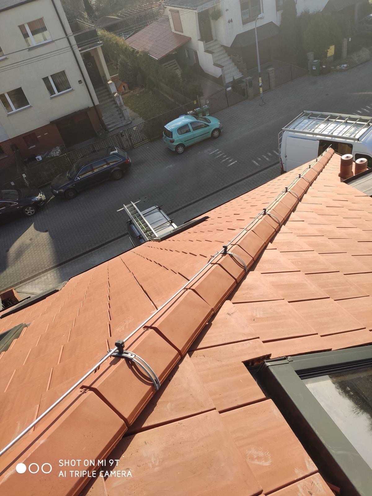 Czerwona dachówka z zamontowanym systemem przewodów, zabezpieczających budynek. W tle widoczny samochód i drabina na dachu furgonetki.