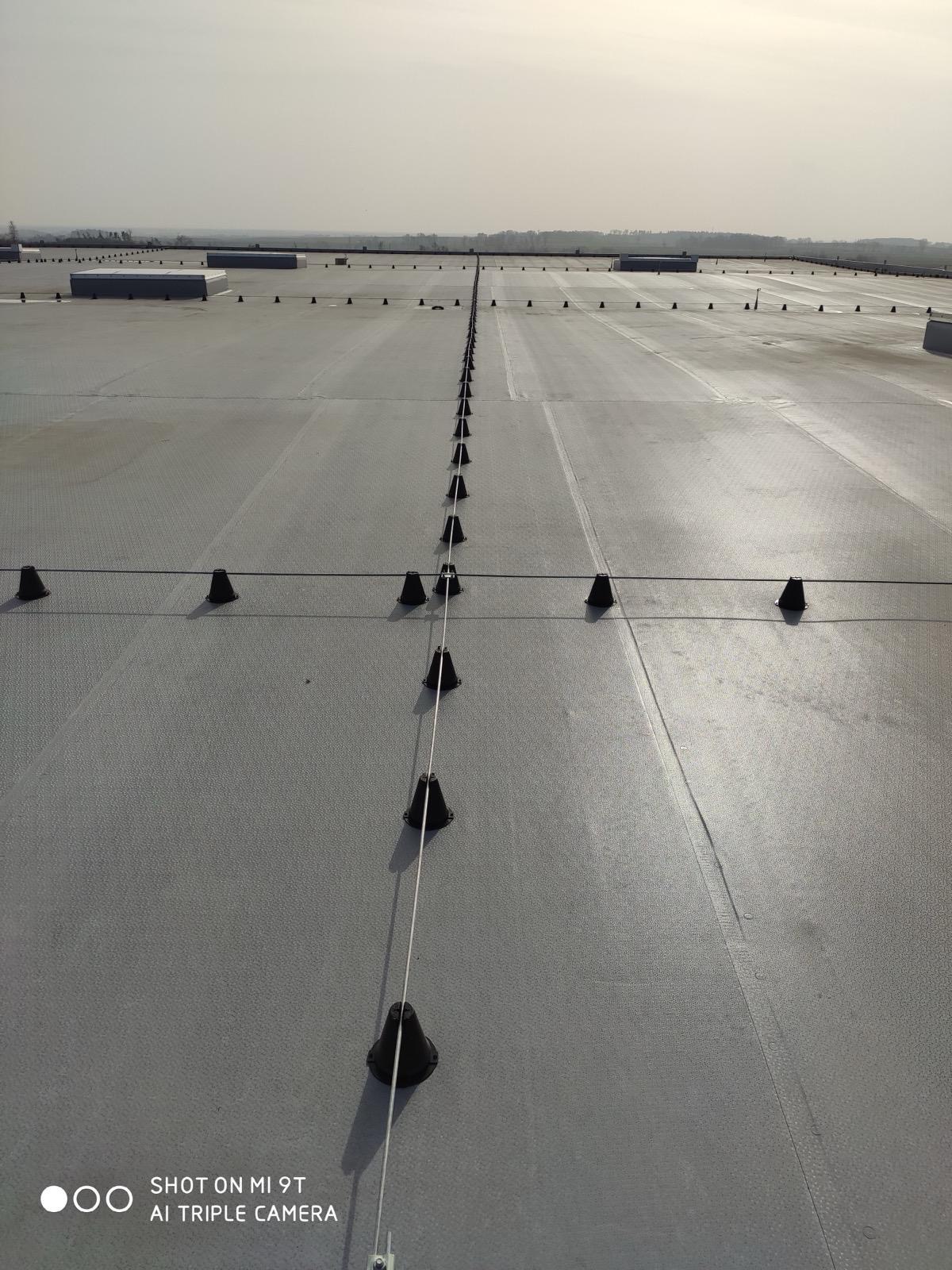 Szary dach przemysłowy z równo rozmieszczonymi, czarnymi uchwytami przewodów. Przewody tworzą siatkę na dachu. Widok z góry, w tle zabudowania.