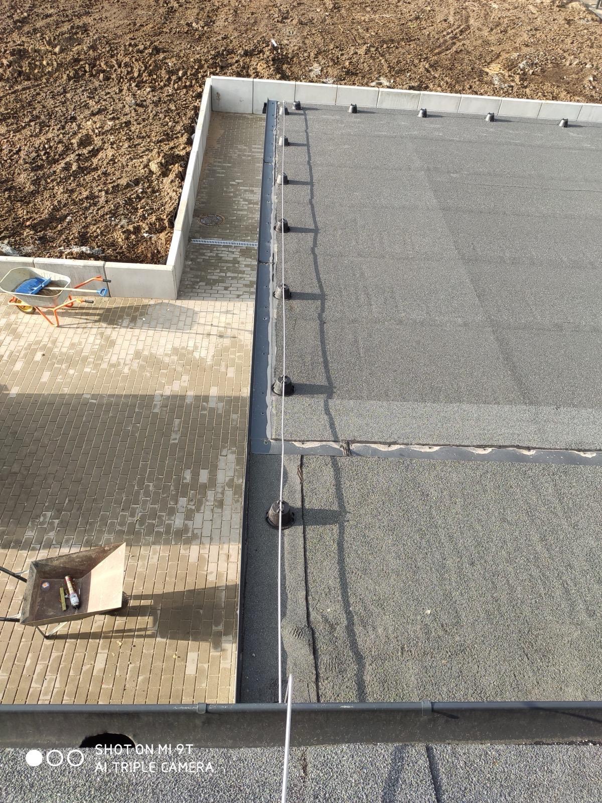 Płaski dach z zamontowanymi uchwytami i przewodami. Widoczne elementy systemu na tle kostki brukowej i ziemi. Prace na ukończeniu, widoczny wózek z narzędziami.