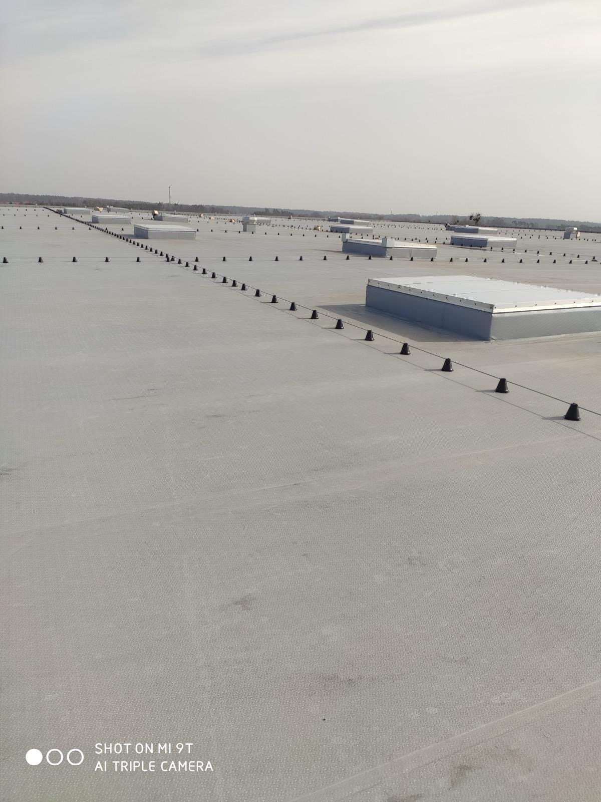 Rozległy dach przemysłowy z systemem ochrony, czarne uchwyty i przewody tworzą regularny wzór, widoczne świetliki dachowe. Daleki horyzont z zielenią.