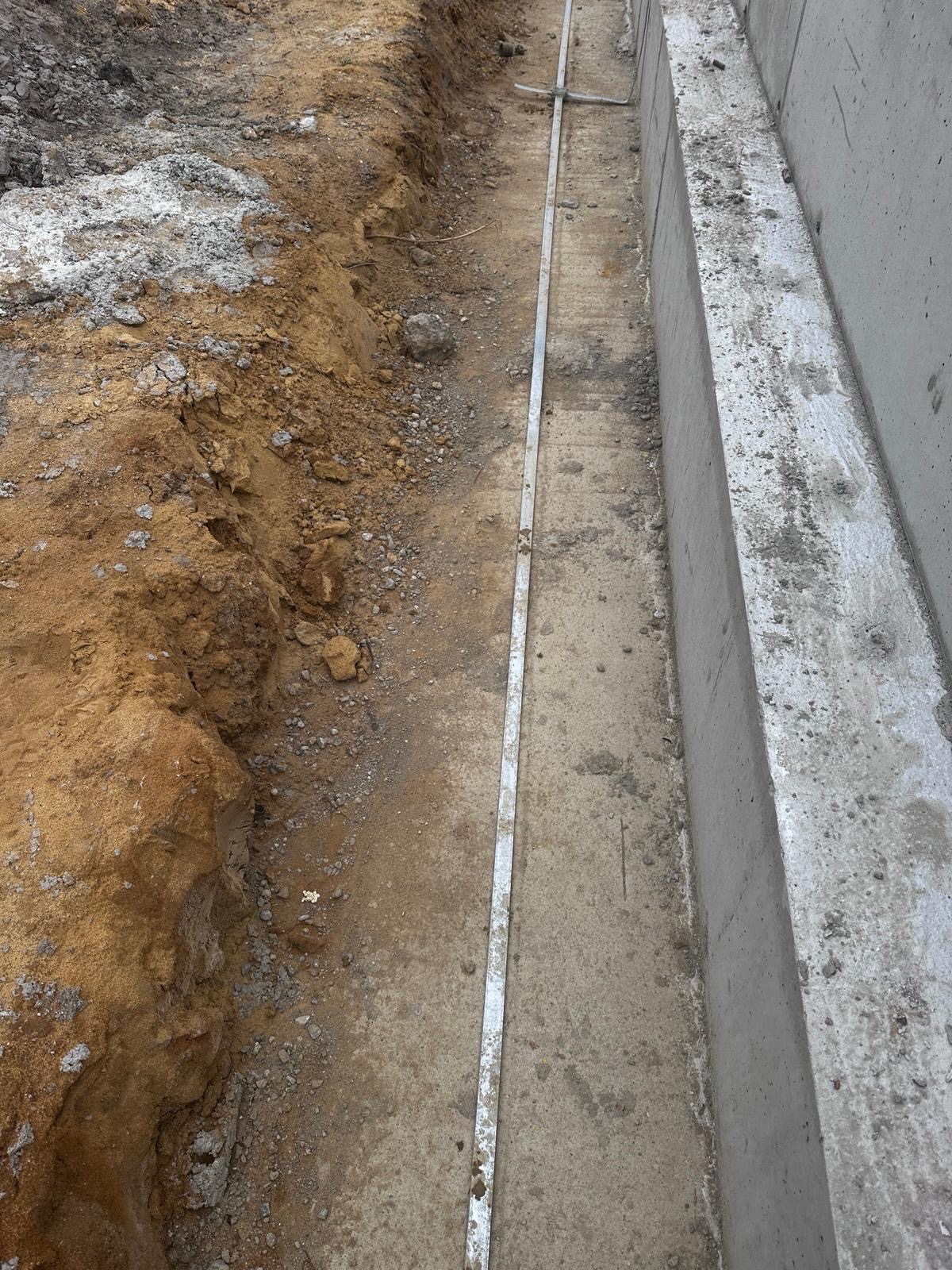 Wykop z ułożonym bednarką przy betonowym fundamencie. Widoczna instalacja uziemiająca w ziemi. Kątowe ujęcie pokazuje detale montażu i przygotowanie do zasypania.