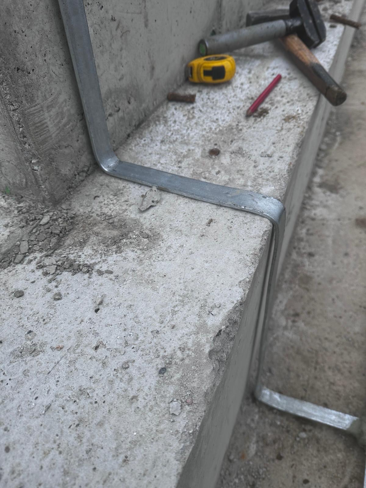 Mocowanie metalowej bednarki do betonowego fundamentu. W tle narzędzia: młotek, miarka, ołówek. Element instalacji odgromowej w trakcie montażu.