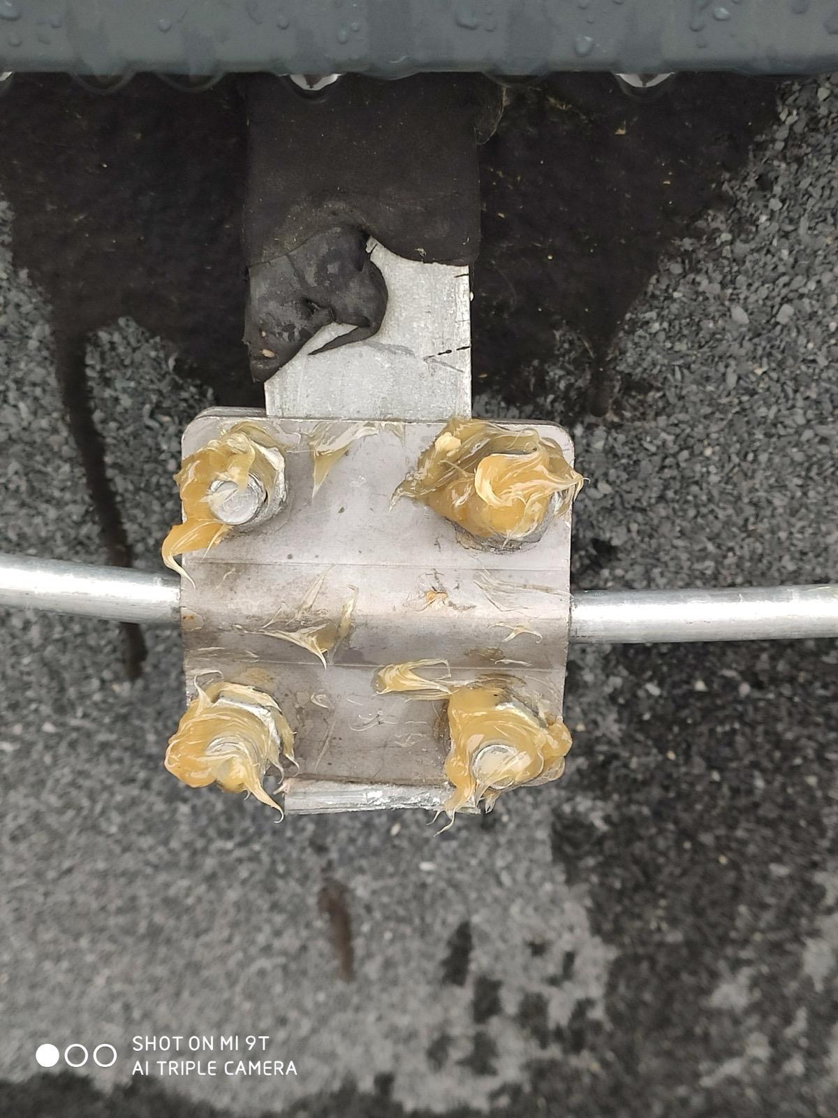 Metalowy uchwyt z nałożonym smarem na śruby, element instalacji zabezpieczającej przed wyładowaniami atmosferycznymi, widok z dołu na tle szarego podłoża.