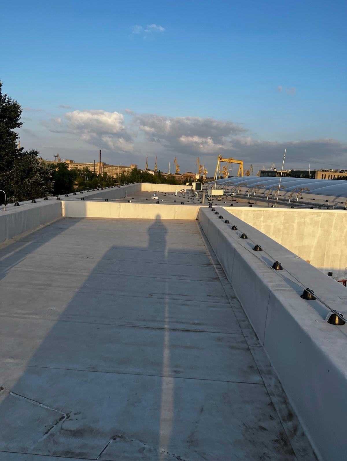 Płaski dach budynku z zainstalowanymi elementami systemu ochrony odgromowej; widoczny cień osoby, w tle zabudowania przemysłowe i niebo.