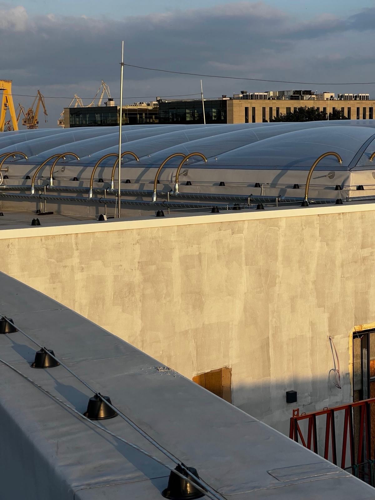 System ochrony odgromowej na dachu budynku z elementami w kolorze złotym, widok z góry, w tle budynki i dźwigi portowe.