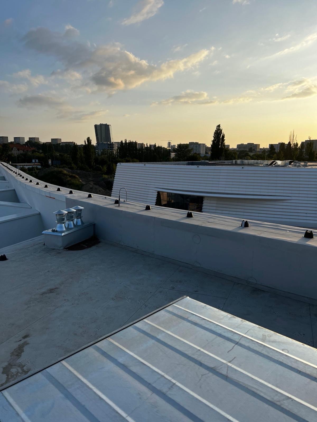 Metalowy dach budynku z elementami instalacji, widok na miasto w tle pod zachmurzonym niebem o zachodzie słońca.