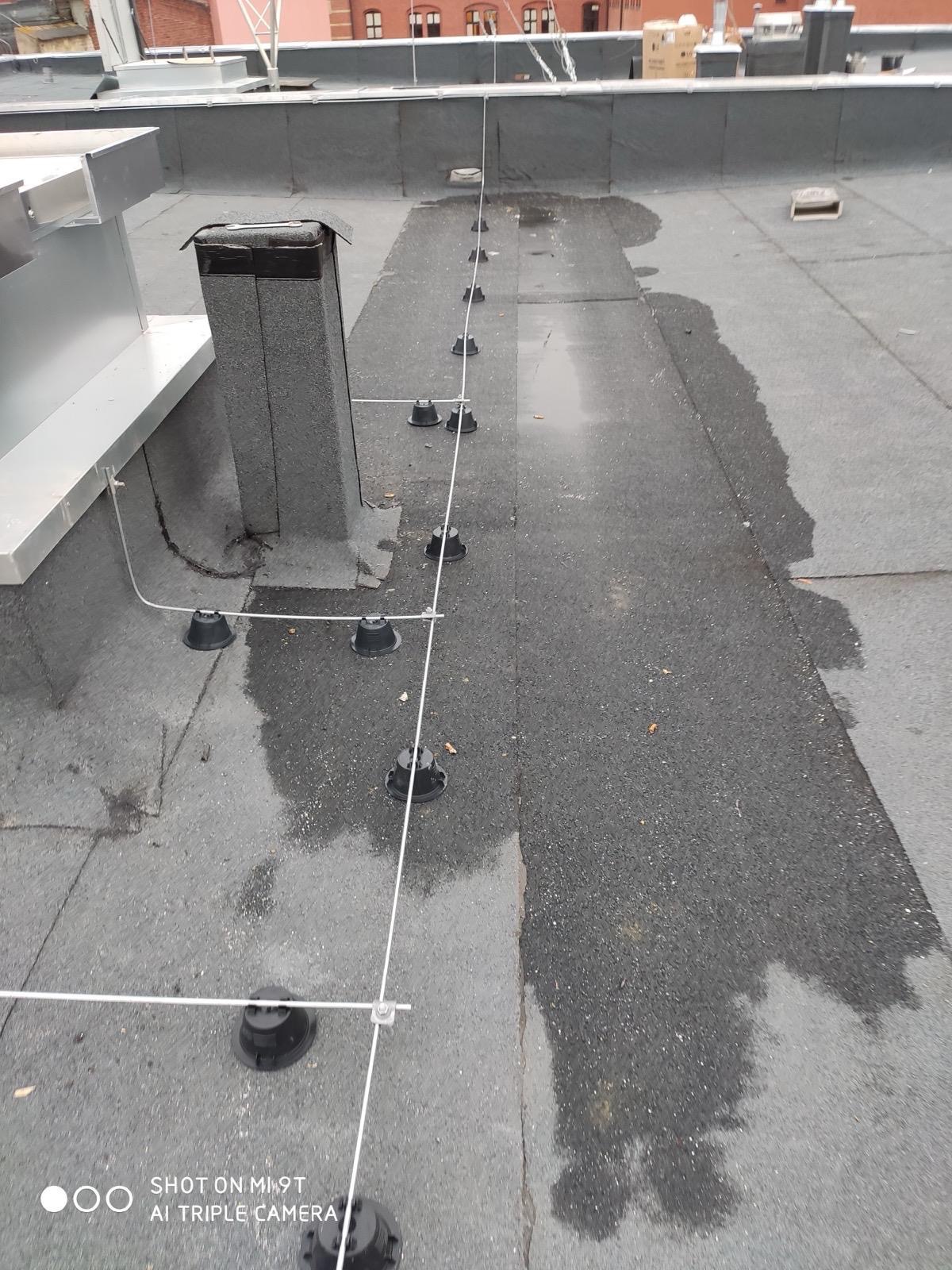 Montaż instalacji odgromowej na płaskim dachu pokrytym papą. Widoczne czarne uchwyty i przewody odgromowe, fragment komina. Część dachu mokra po deszczu.