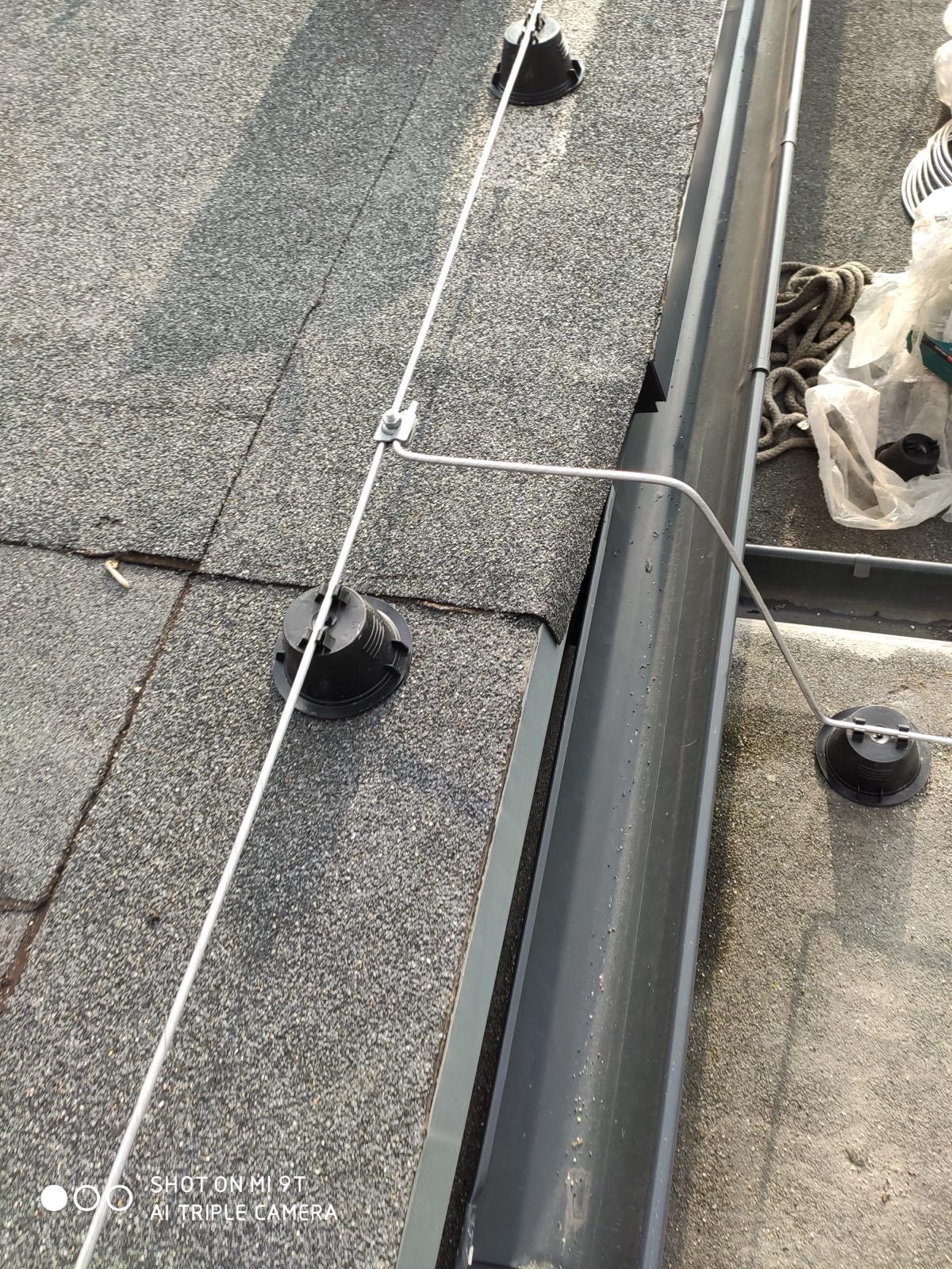 Szara dachówka z elementami systemu zabezpieczenia przed wyładowaniami atmosferycznymi: uchwyty, przewód i rynna. Widoczne narzędzia pracy i lina.