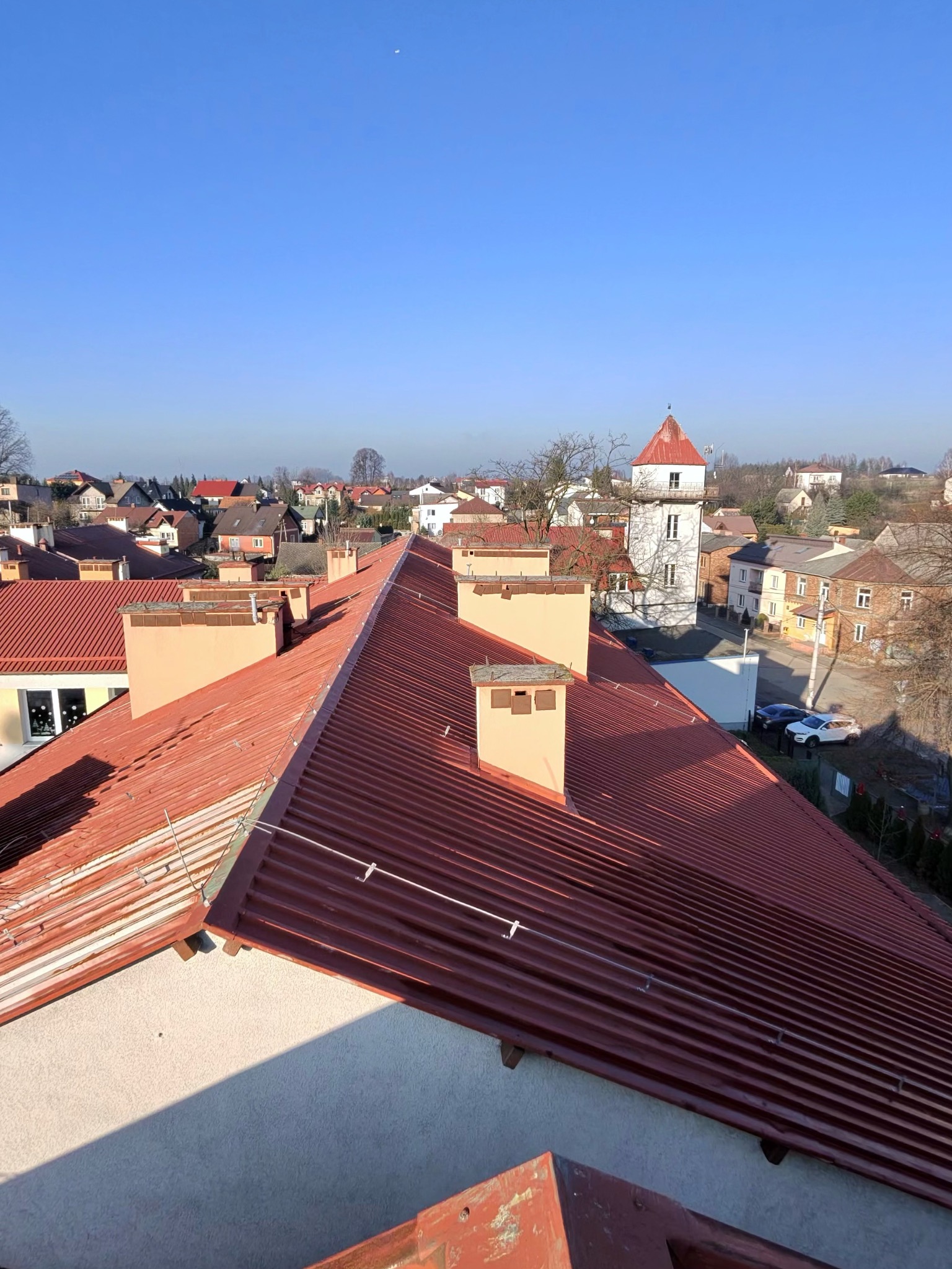 Czerwony dach z blachy trapezowej z zamontowanym systemem ochrony odgromowej na tle zabudowy miejskiej i błękitnego nieba.