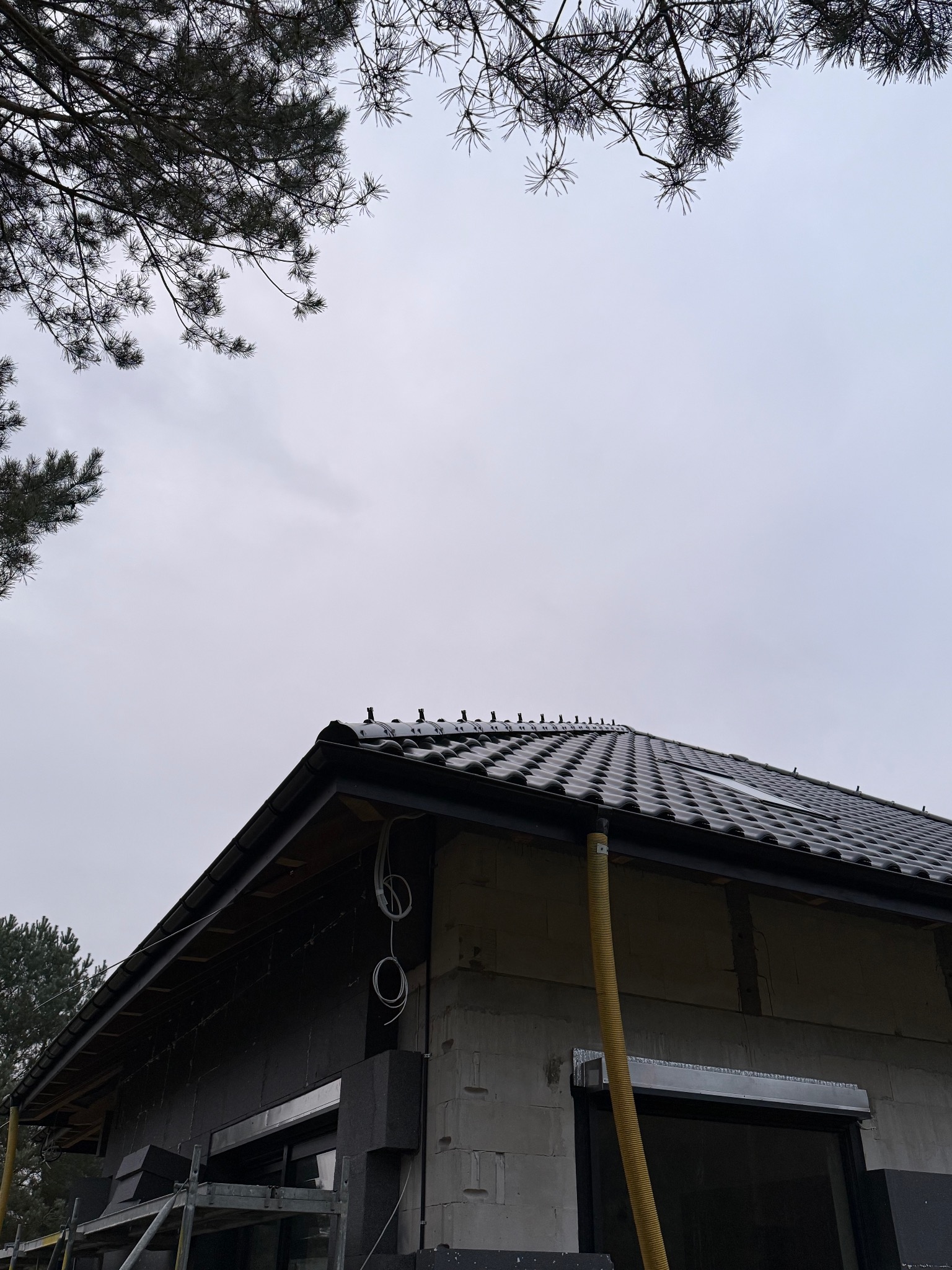 Dom w trakcie budowy z zamontowaną instalacją odgromową na dachu z czarnej dachówki. Widoczne przewody i rusztowanie. Ujęcie z dołu, pochmurne niebo.