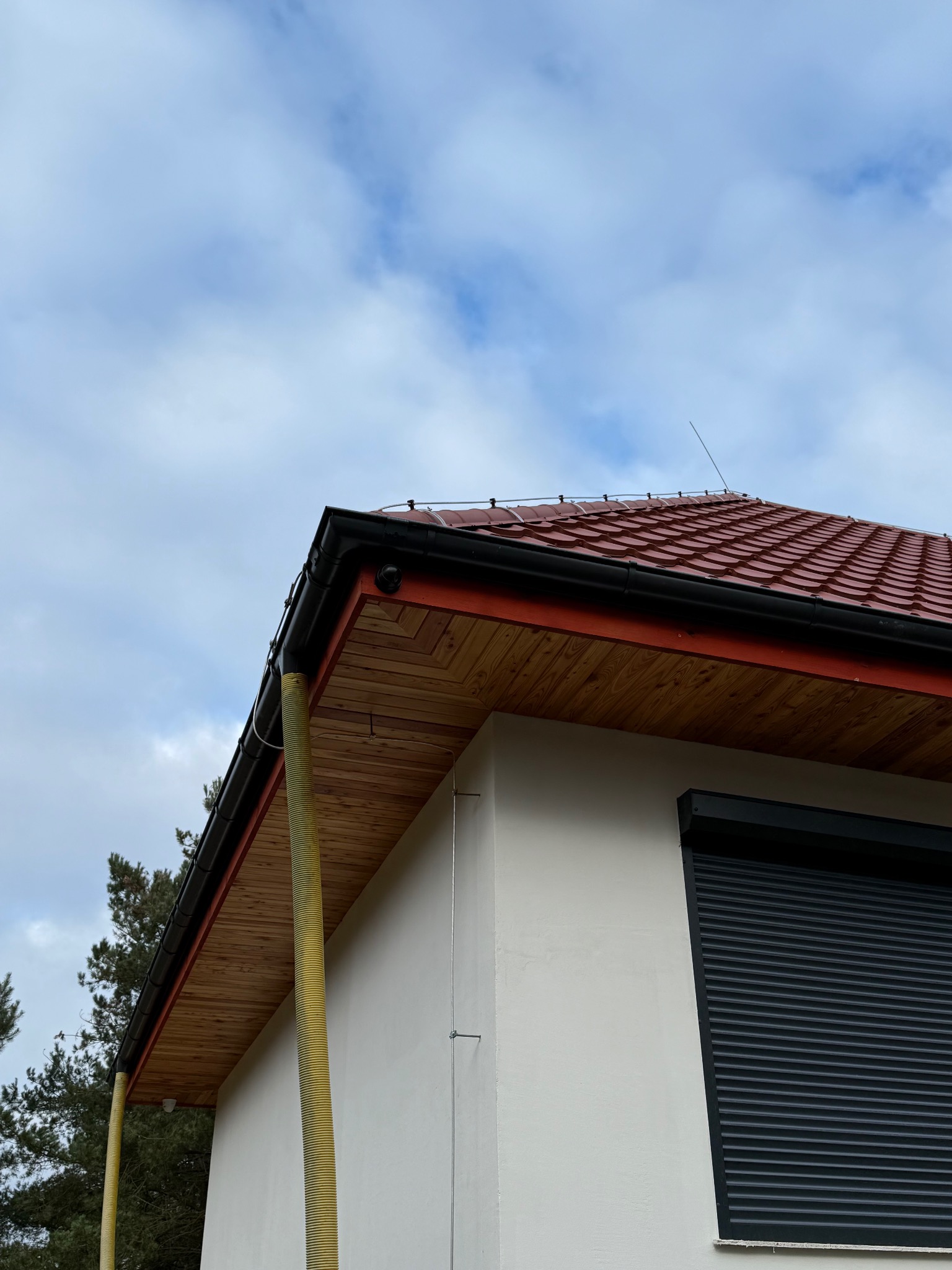 Fragment domu z czerwoną dachówką, rynnami i elementami systemu ochrony odgromowej, widoczne z dołu na tle pochmurnego nieba.
