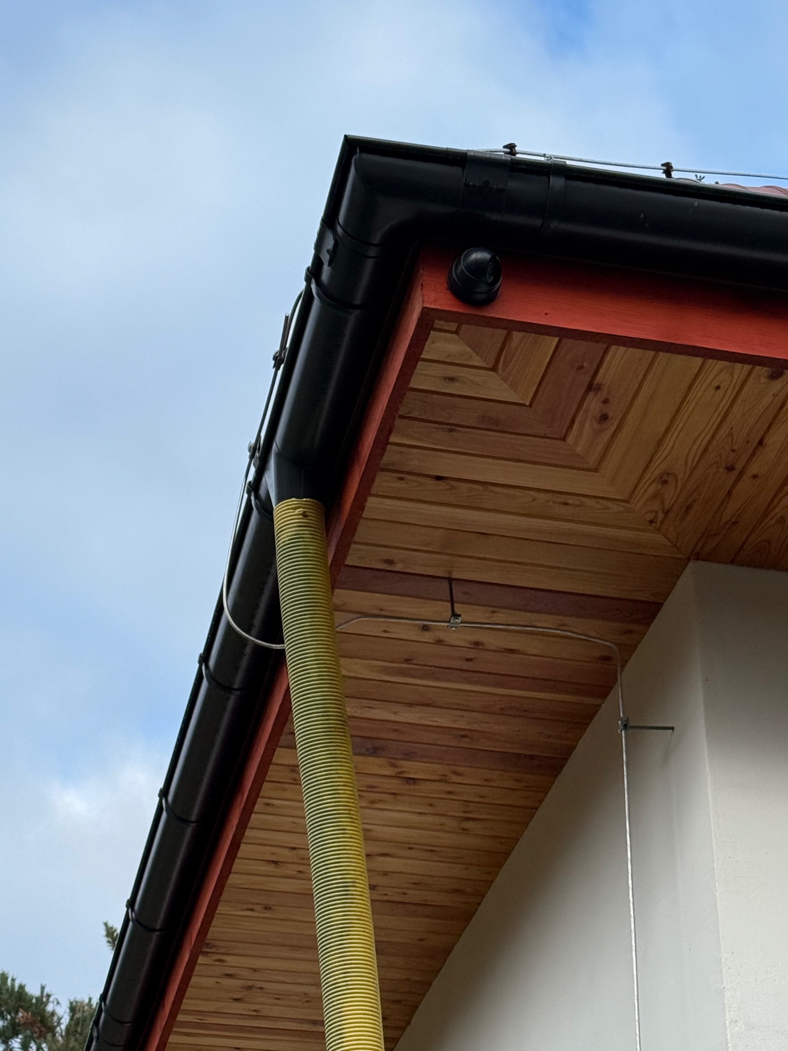 Fragment dachu z czarną rynną i żółtą rurą spustową, widok z dołu na tle jasnego nieba. Widoczna drewniana podbitka i element instalacji na ścianie budynku.