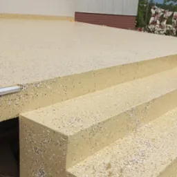 Beżowe schody zewnętrzne z posadzką żywiczną w plamki, prowadzące do budynku. Widoczne elementy wykończenia i fragment tła z zielenią.