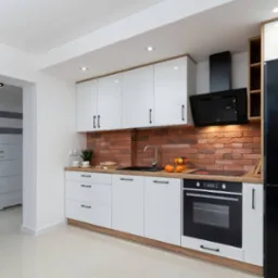 Nowoczesna kuchnia z białymi szafkami, ceglaną ścianą i czarnym sprzętem AGD. Jasne płytki podłogowe i oświetlenie punktowe. Widok na otwarte przejście do innego pomieszczenia.