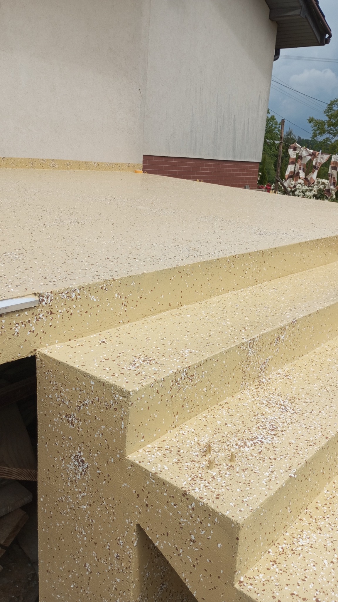 Beżowe schody zewnętrzne z posadzką żywiczną w plamki, prowadzące do budynku. Widoczne elementy wykończenia i fragment tła z zielenią.