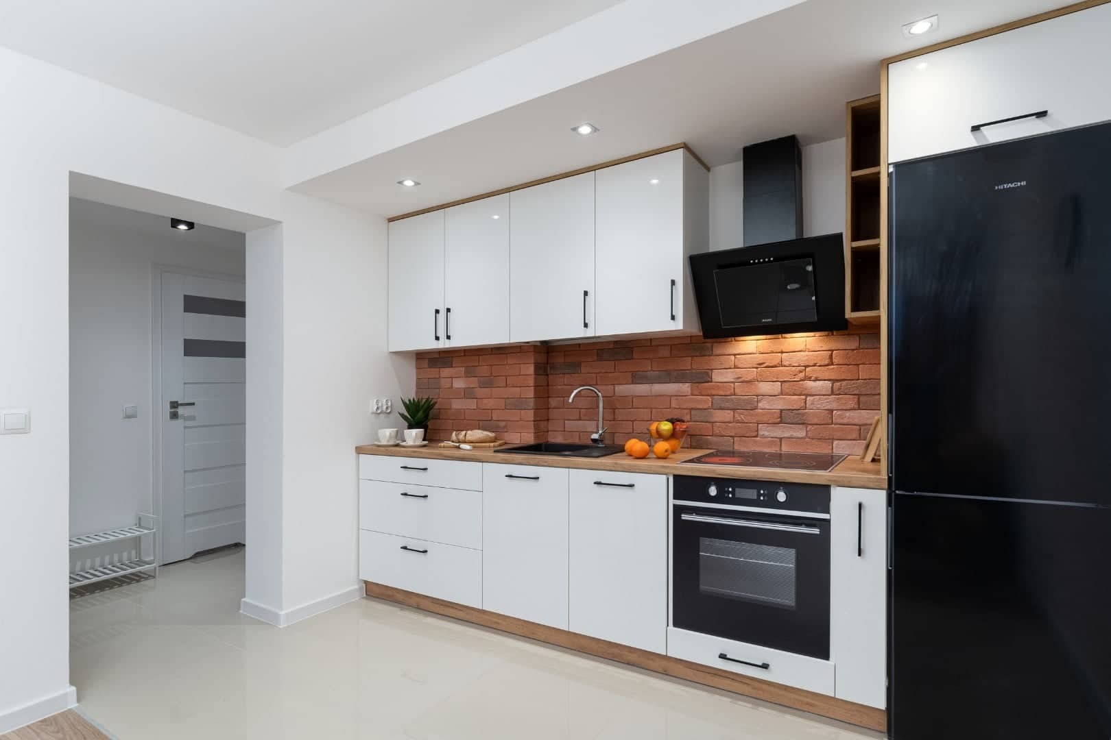Nowoczesna kuchnia z białymi szafkami, ceglaną ścianą i czarnym sprzętem AGD. Jasne płytki podłogowe i oświetlenie punktowe. Widok na otwarte przejście do innego pomieszczenia.