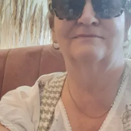 Portret kobiety w okularach przeciwsłonecznych, w tle dekoracyjne trawy i element wnętrza. Ubrana w bluzkę z haftowanymi kwiatami.