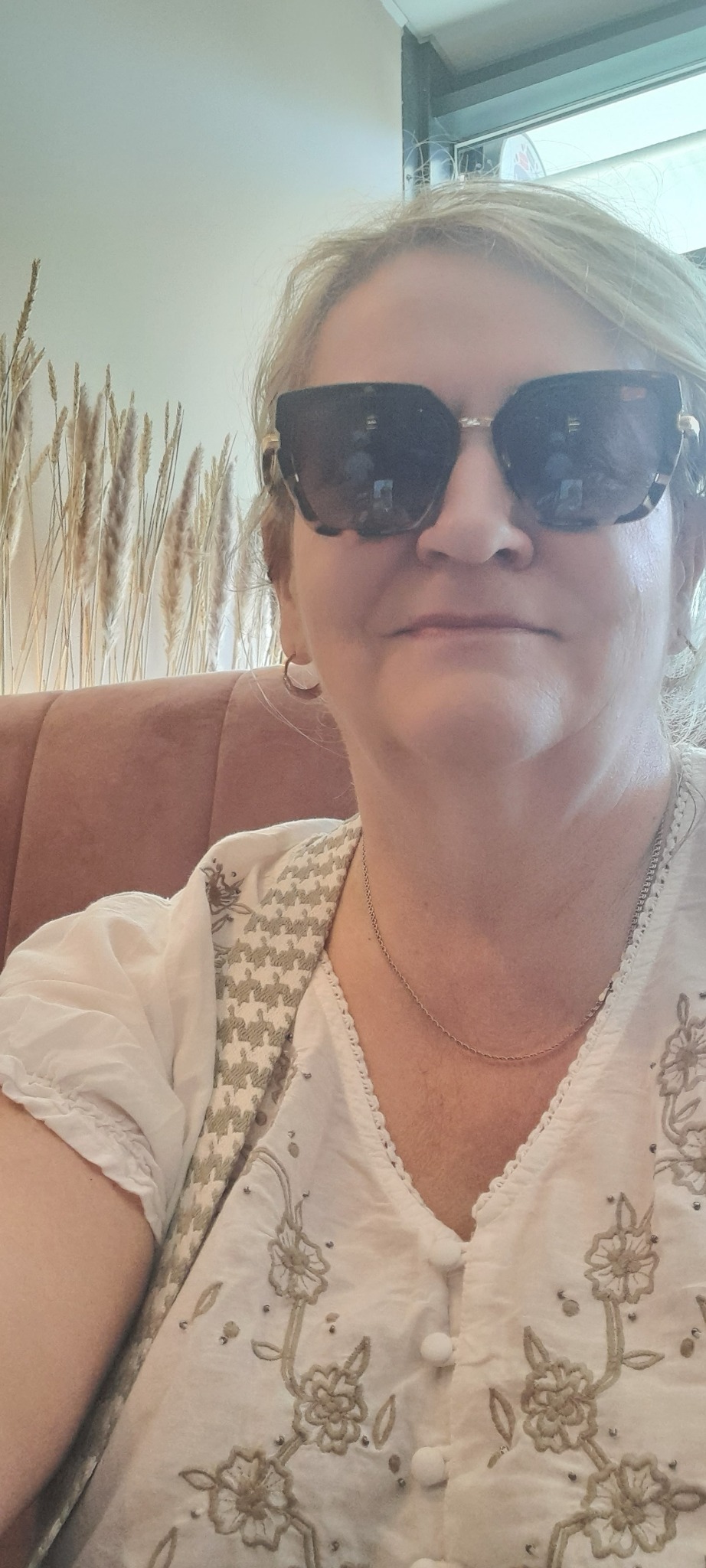 Portret kobiety w okularach przeciwsłonecznych, w tle dekoracyjne trawy i element wnętrza. Ubrana w bluzkę z haftowanymi kwiatami.