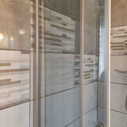 Małgorzata Aneta - Szklana kabina prysznicowa z chromowaną armaturą, obłożona mozaiką i płytkami w odcieniach beżu i szarości. Widoczne detale wykończenia i czystość powierzchni.