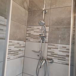 Małgorzata Aneta - Nowoczesna kabina prysznicowa z deszczownicą i baterią, wykończona beżowymi płytkami z dekoracyjnym pasem mozaiki. Widoczny fragment szklanej ścianki kabiny.