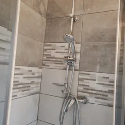 Nowoczesna kabina prysznicowa z deszczownicą i baterią, wykończona beżowymi płytkami z dekoracyjnym pasem mozaiki. Widoczny fragment szklanej ścianki kabiny.