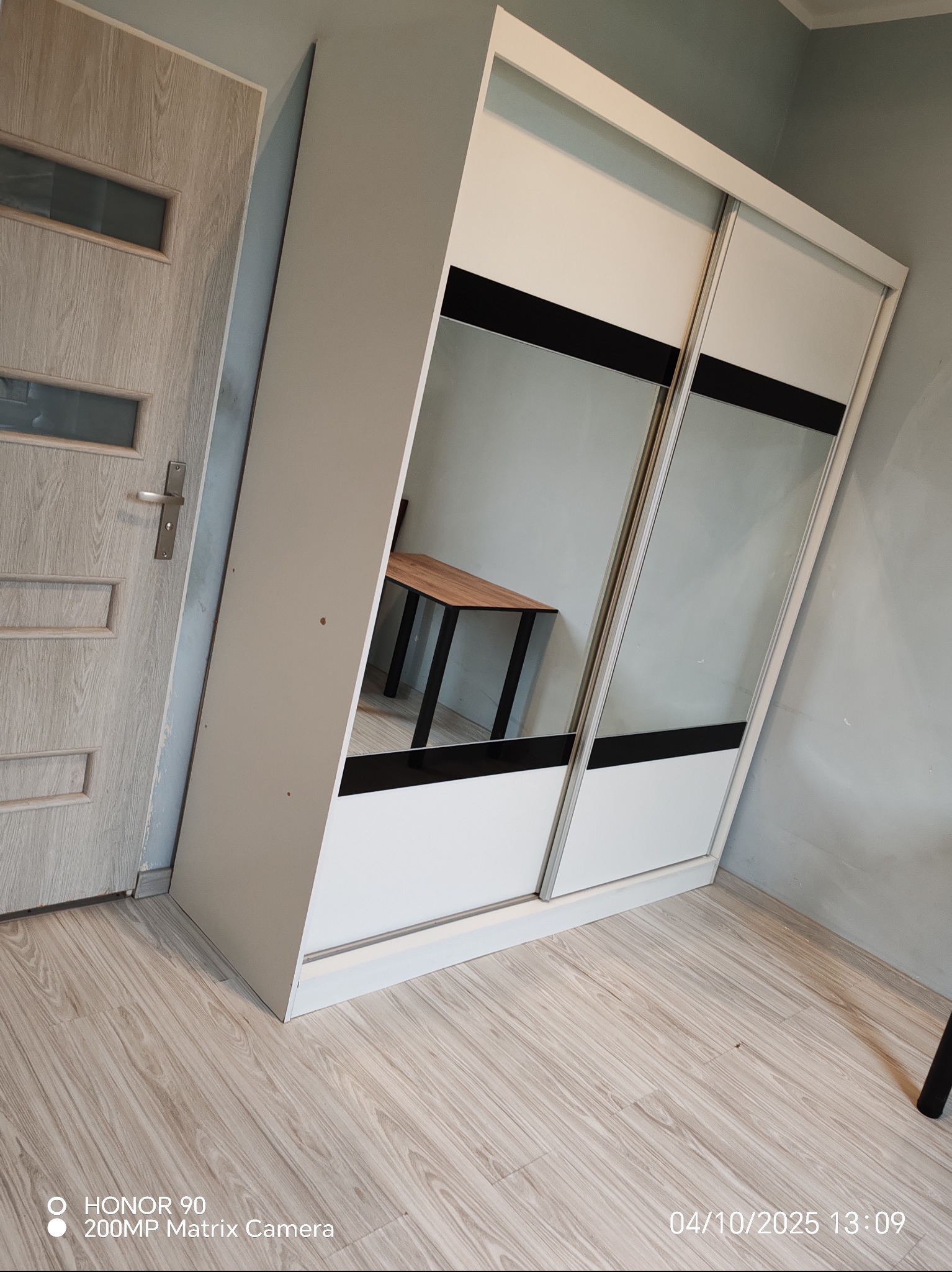 Biała szafa przesuwna z lustrami i czarnymi pasami, ustawiona przy ścianie i drzwiach z drewnopodobnym wzorem, na podłodze z paneli. Widoczne odbicie stołu.