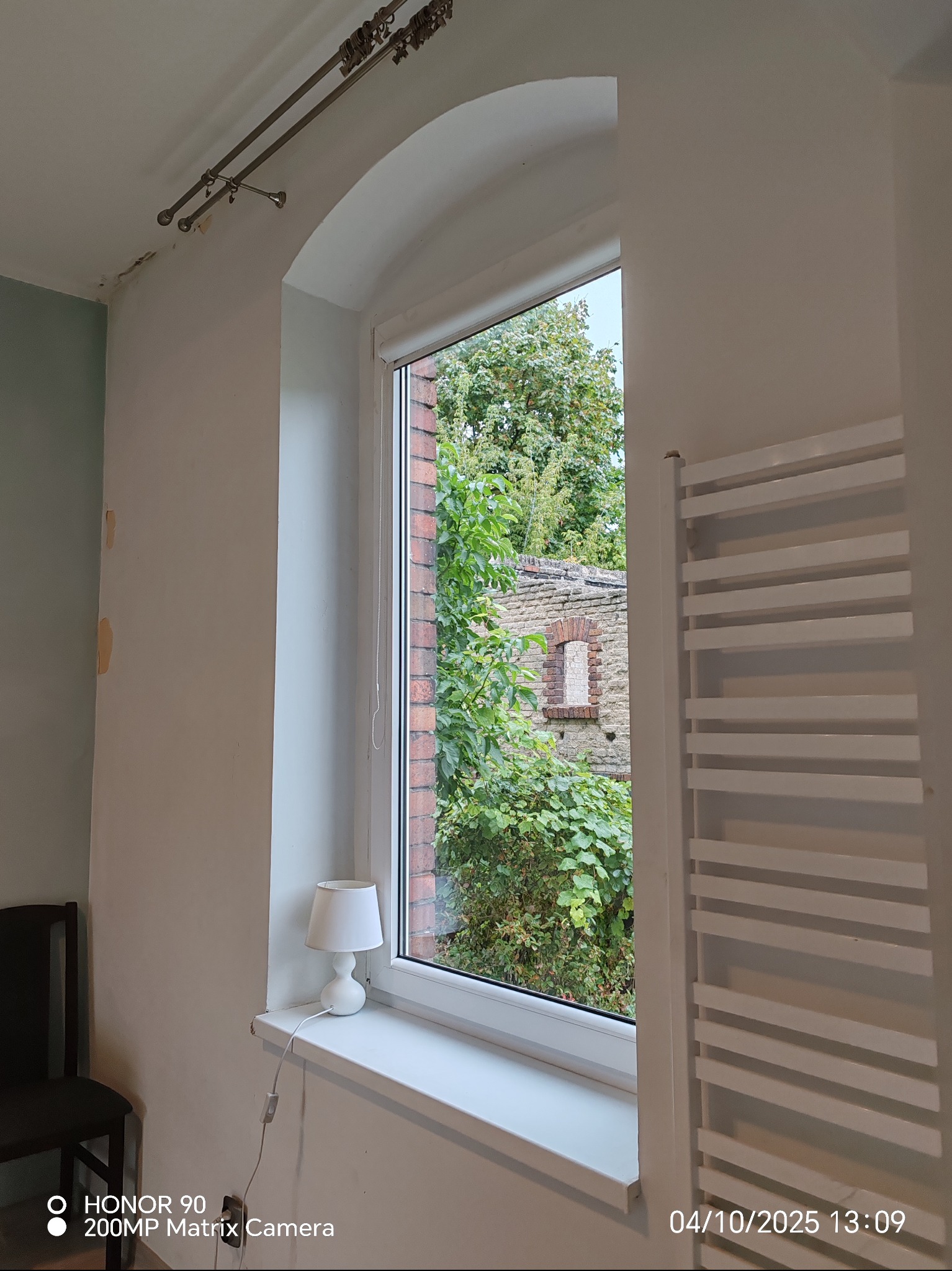 Arched window view: greenery, brick wall. Interior: radiator, lamp on sill, peeling paint near curtain rod. Natural light, residential setting.