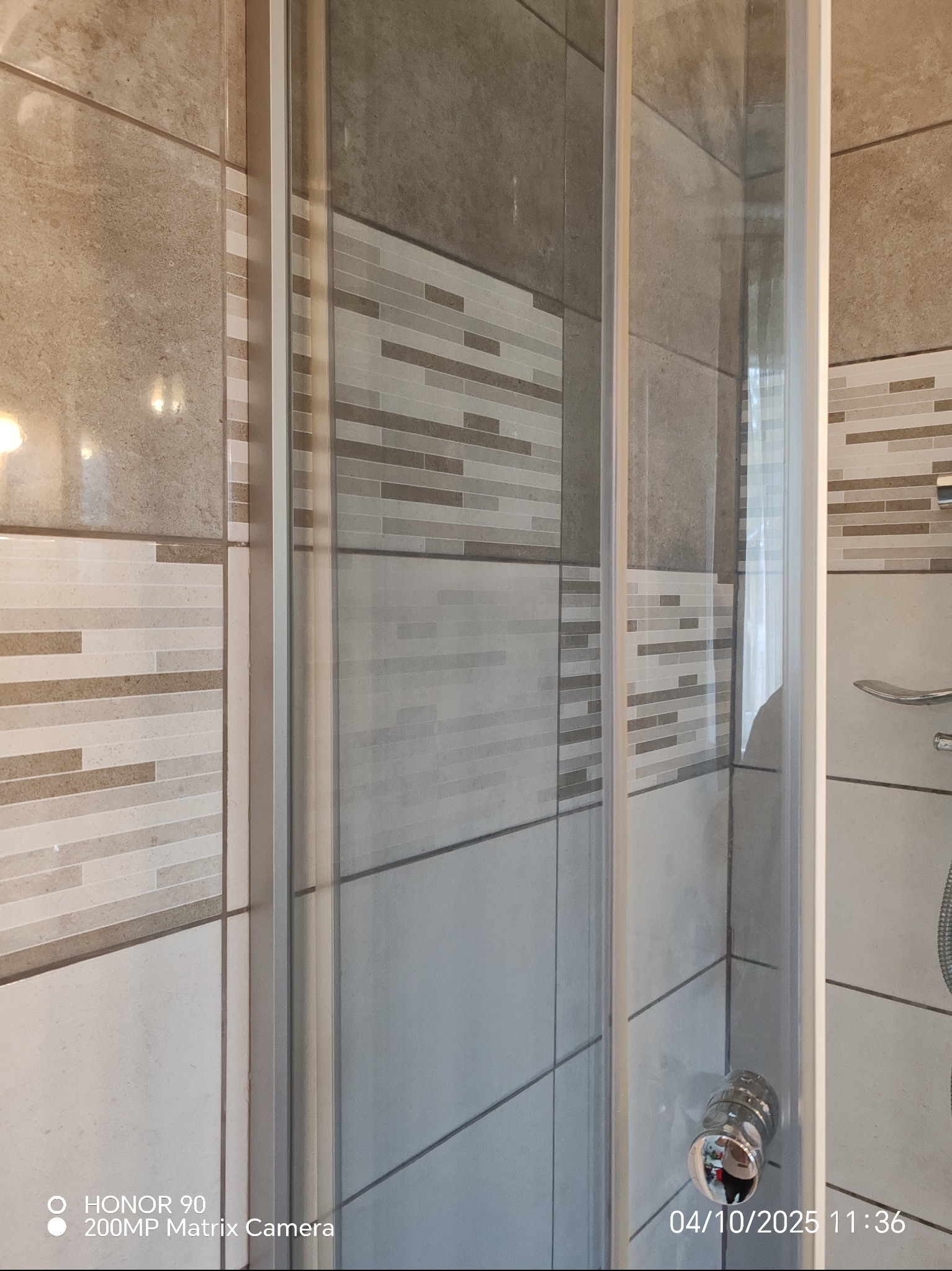 Szklana kabina prysznicowa z chromowaną armaturą, obłożona mozaiką i płytkami w odcieniach beżu i szarości. Widoczne detale wykończenia i czystość powierzchni.