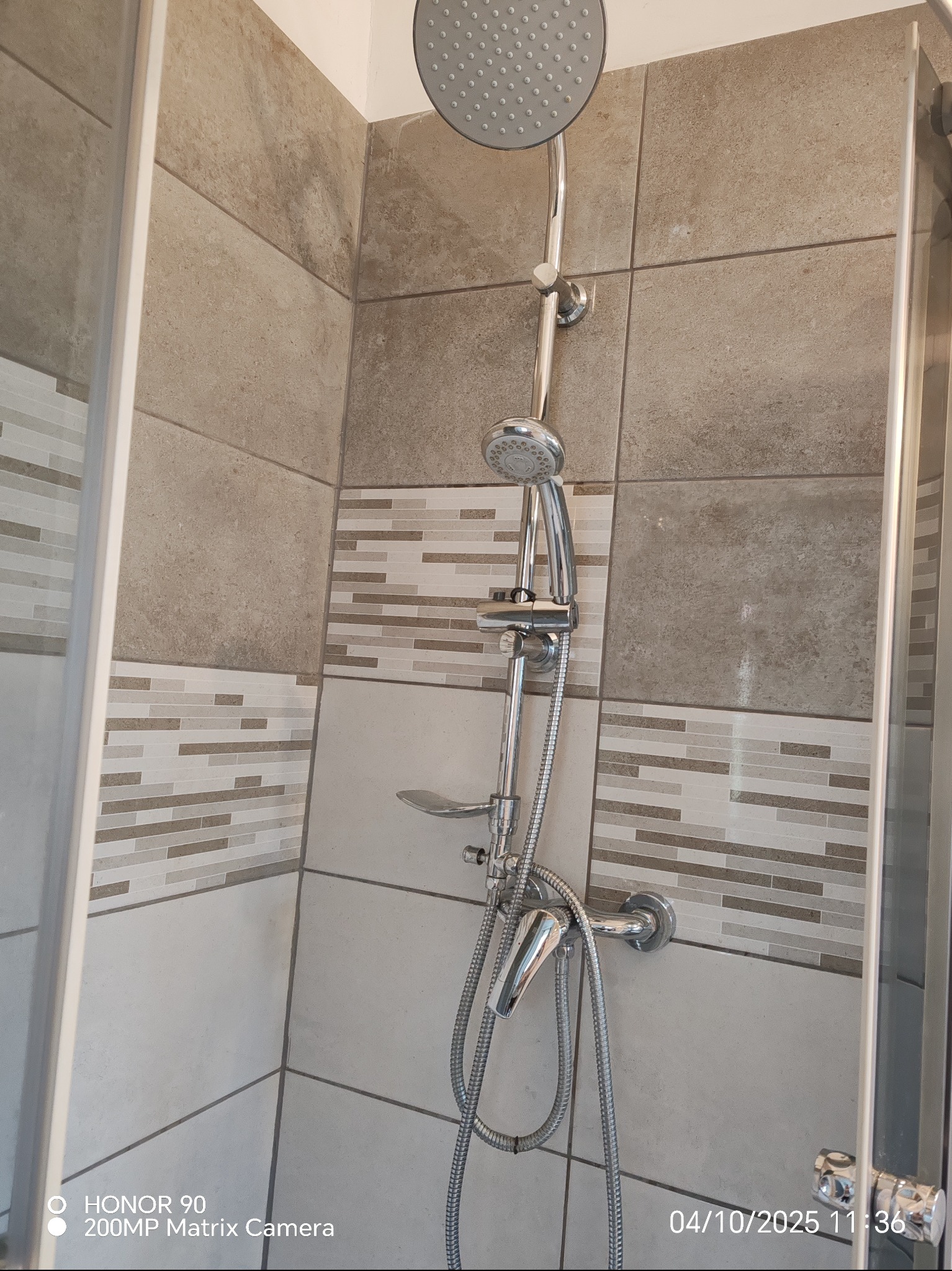 Nowoczesna kabina prysznicowa z deszczownicą i baterią, wykończona beżowymi płytkami z dekoracyjnym pasem mozaiki. Widoczny fragment szklanej ścianki kabiny.