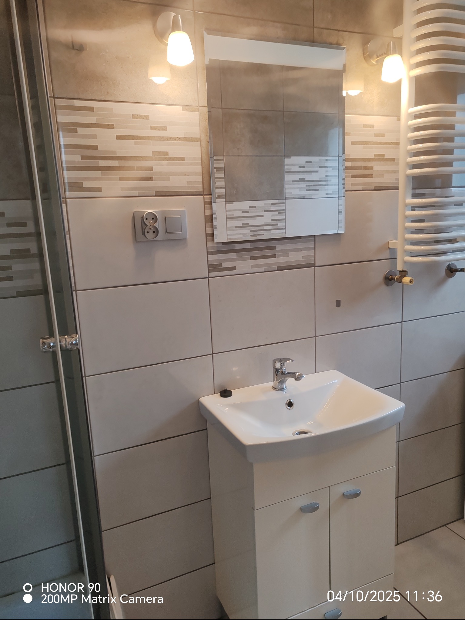 Umywalka z szafką w łazience z mozaiką, grzejnikiem drabinkowym i oświetleniem punktowym. Widoczny fragment kabiny prysznicowej.